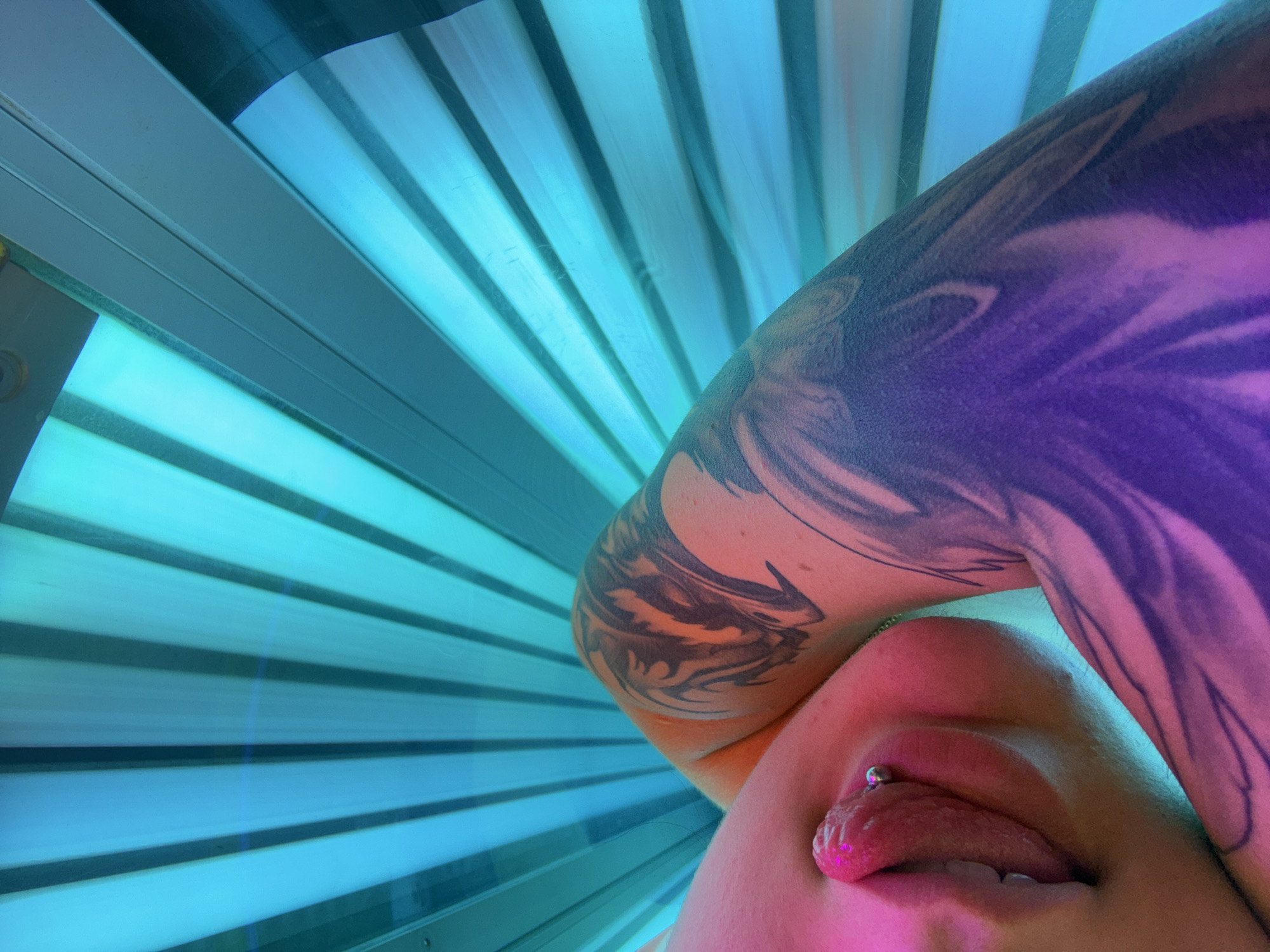 The image shows a close-up view of a person's face and arm, illuminated by the blue and pink lights of a tanning bed. The person's left arm, adorned with a large black tattoo, is bent upwards, partially obscuring their face. Their face is tilted, with their lips slightly parted, revealing their tongue, which has a silver stud piercing. The background is a series of blue and white horizontal lines, characteristic of a tanning bed's interior. The lighting creates a strong contrast between the illuminated skin and the darker tattoo. The overall mood is artificial and intimate, likely captured during a private tanning session. No visible text is present in the image.