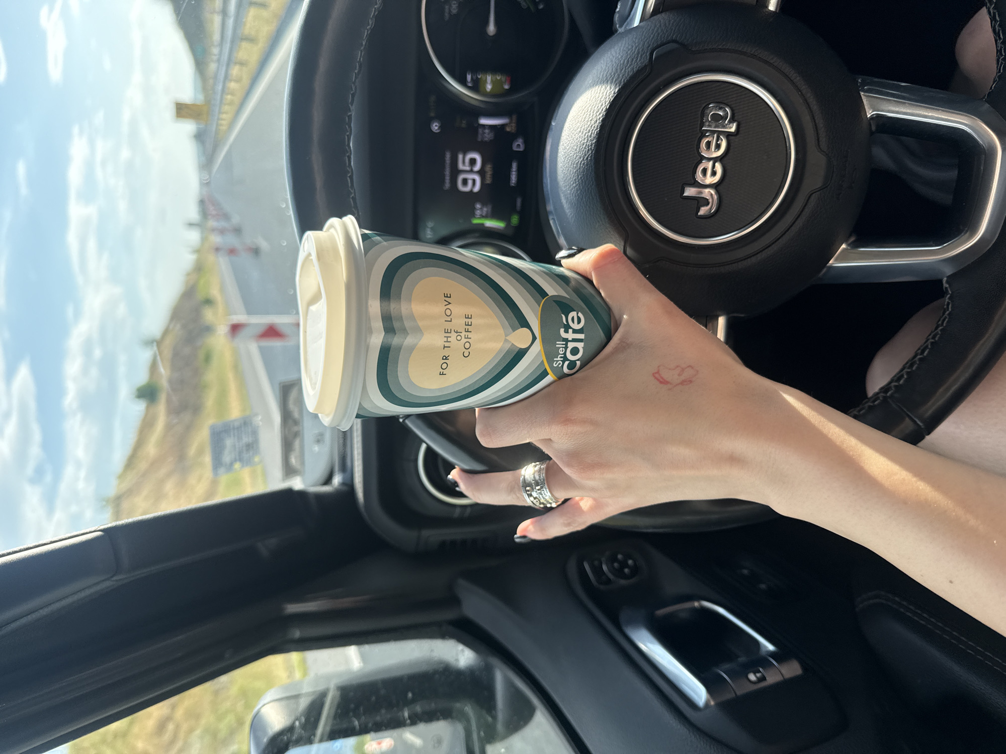A photograph shows the interior of a Jeep vehicle, focusing on the driver's side. The driver's hand is holding a "Shell café" coffee cup with the slogan "FOR THE LOVE OF COFFEE." A ring is visible on the driver's finger, and a small, red tattoo is on their wrist. The Jeep's steering wheel, displaying the "Jeep" logo, is visible. The digital dashboard shows the vehicle traveling at 95 km/h. The exterior of the car shows a highway landscape under a bright, slightly cloudy sky. The photograph was taken on the A2 highway near Hadzhidimitrovo, Bulgaria, evidenced by signage and the overall landscape visible through the windshield.