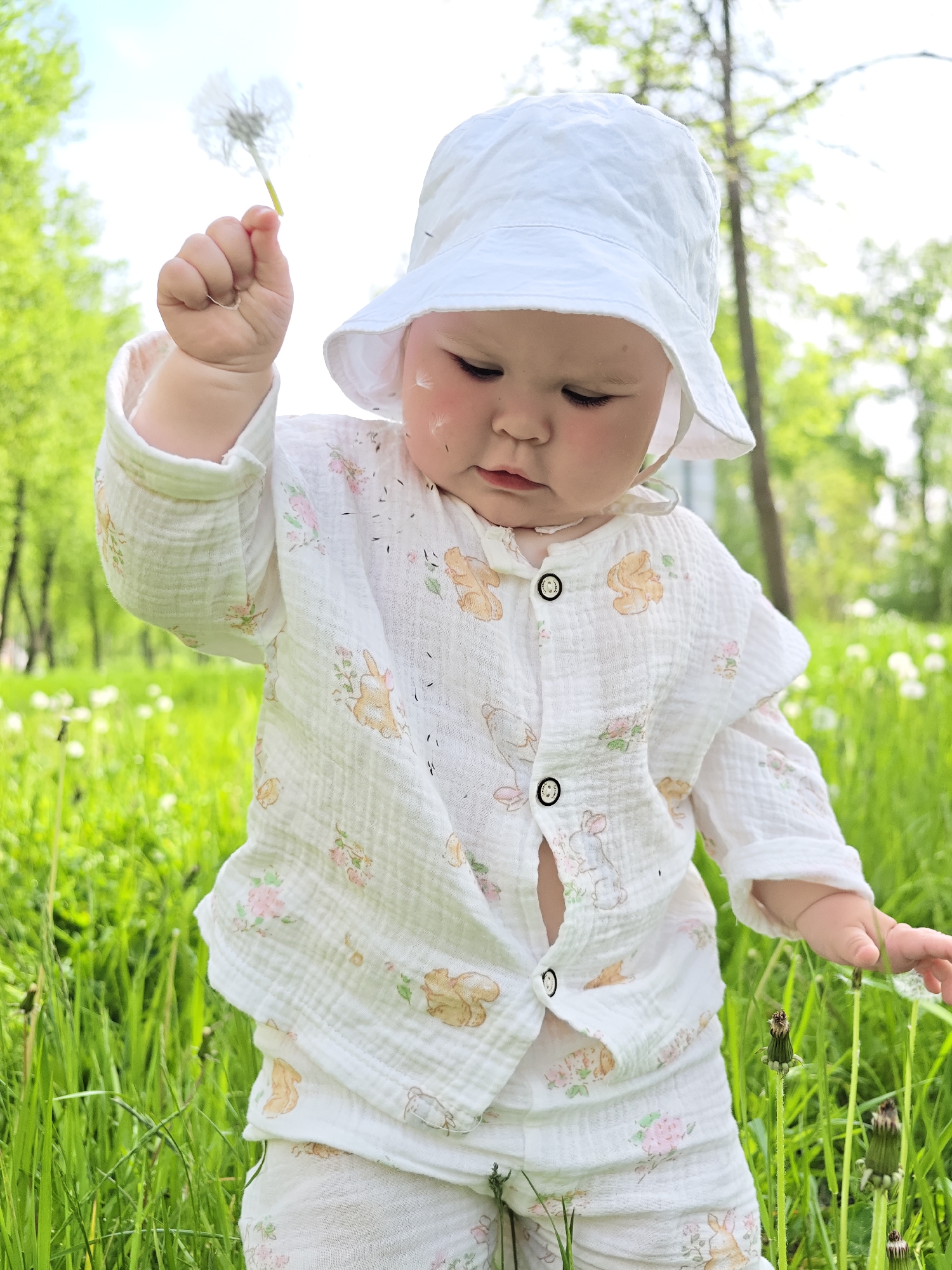 A baby is standing in a grassy field. The baby is wearing a white hat, a white button-up shirt with a bunny and floral pattern, and white pants. The shirt is partially unbuttoned. The baby is holding a dandelion in their left hand and reaching towards another dandelion with their right hand. The background consists of green grass and trees. Dandelion seeds are visible on the baby's face. The location is Chaikovsky, Russia.