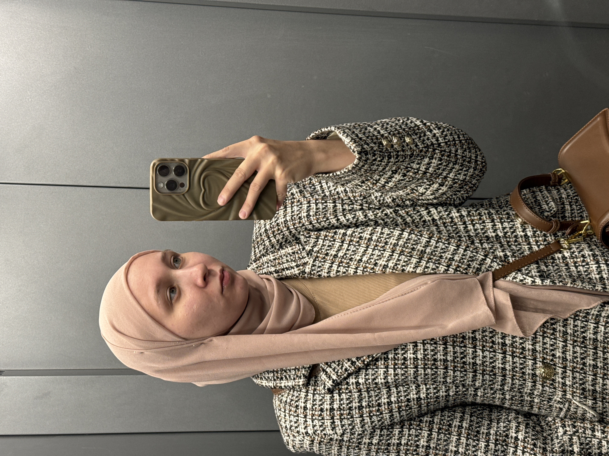 A woman wearing a hijab and a tweed blazer is taking a selfie. She is holding a smartphone with a beige phone case. A brown purse is also visible. The background appears to be a plain, gray wall. The location is not identifiable as Chaikovsky, Russia, from the image provided.