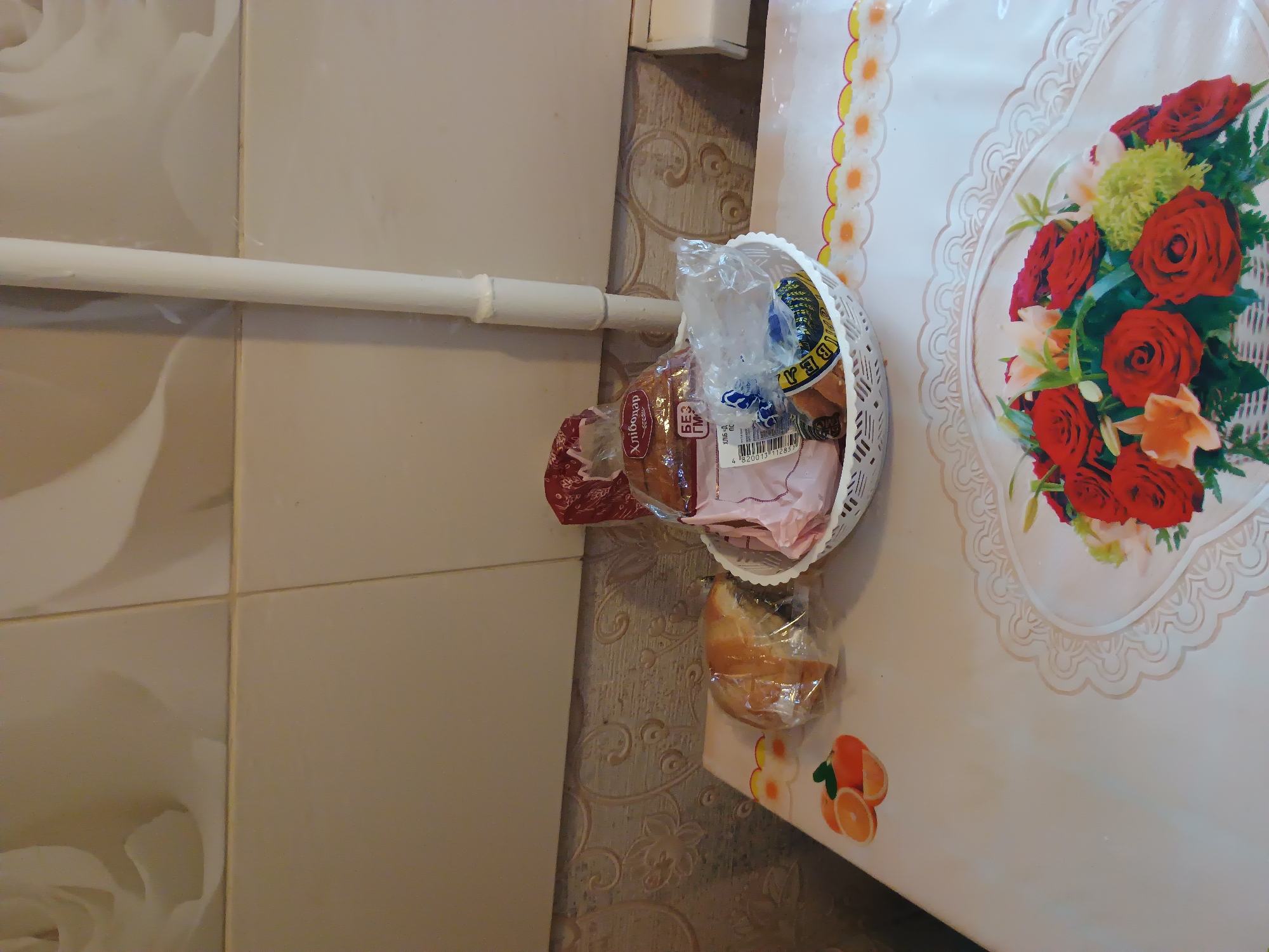 This media file shows a close-up view of a basket filled with baked goods, including loaves of bread and possibly pastries, placed on a table with a floral tablecloth. The basket is white and woven, and it contains several items wrapped in clear plastic. One visible loaf of bread has a label with text in Cyrillic script, which appears to say "Хлібодар". The tablecloth is light-colored with a decorative lace-like border and a prominent illustration of a bouquet of red roses and other flowers. To the right of the basket, on the tablecloth, are stickers or prints of sliced oranges. The background shows a tiled wall and a portion of what appears to be a radiator or pipe. The lighting suggests an indoor setting, likely a kitchen or dining area. The scene is still and does not depict any people or ongoing activity. The overall impression is of a domestic setting in Ukraine, possibly Zaporizhzhia, given the provided location context. There are no discernible indicators of the time of day or weather conditions.