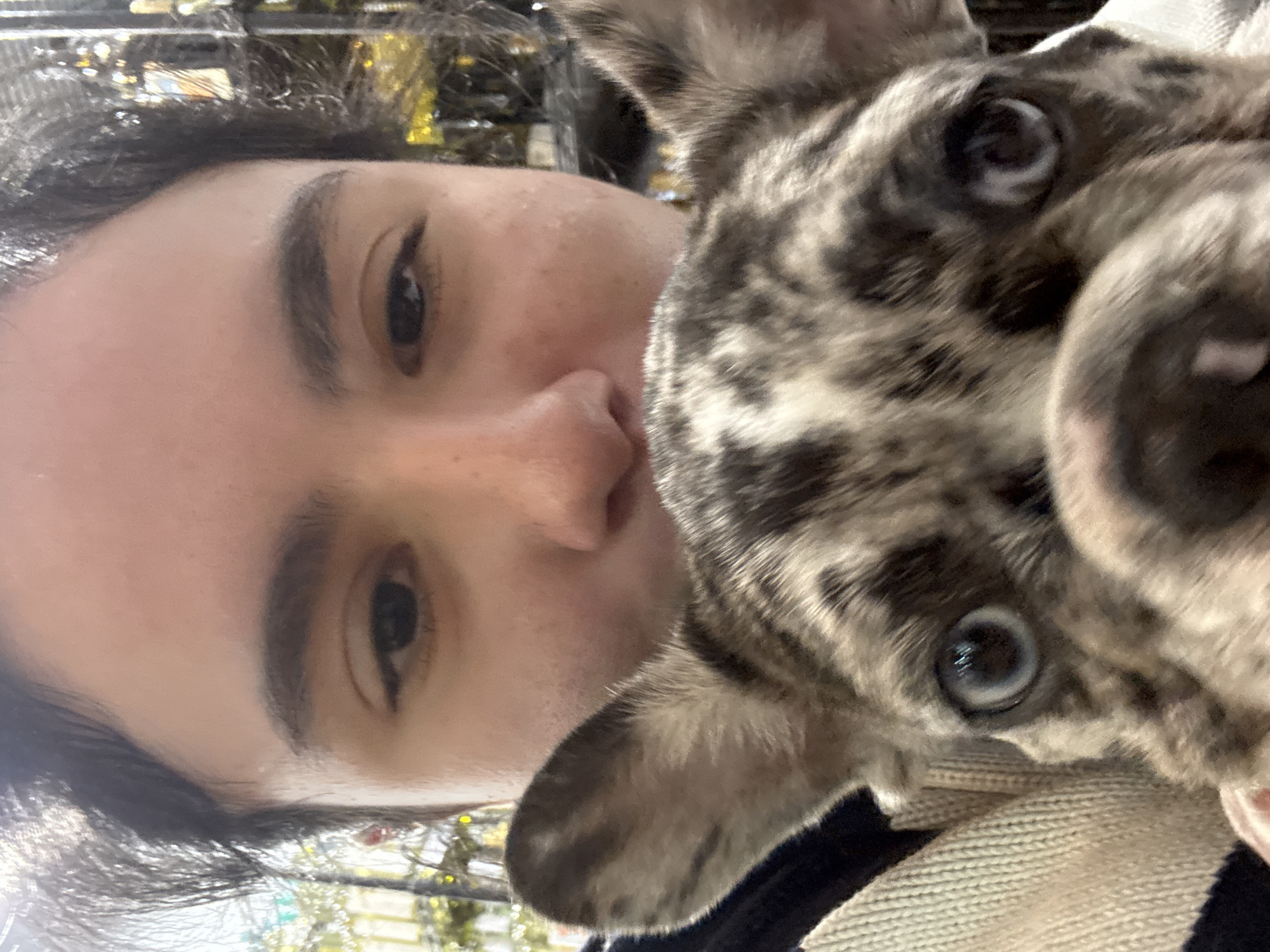 A close-up, slightly blurred selfie features a person with dark hair and brown eyes positioned on the left. The person's face is partially obscured by a brindle-patterned dog with light-colored eyes pressed against their face on the right. The dog's nose and mouth are close to the camera. The background shows a blurred, indistinct cityscape with hints of greenery and a metallic object. The location is possibly Austin, United States of America.