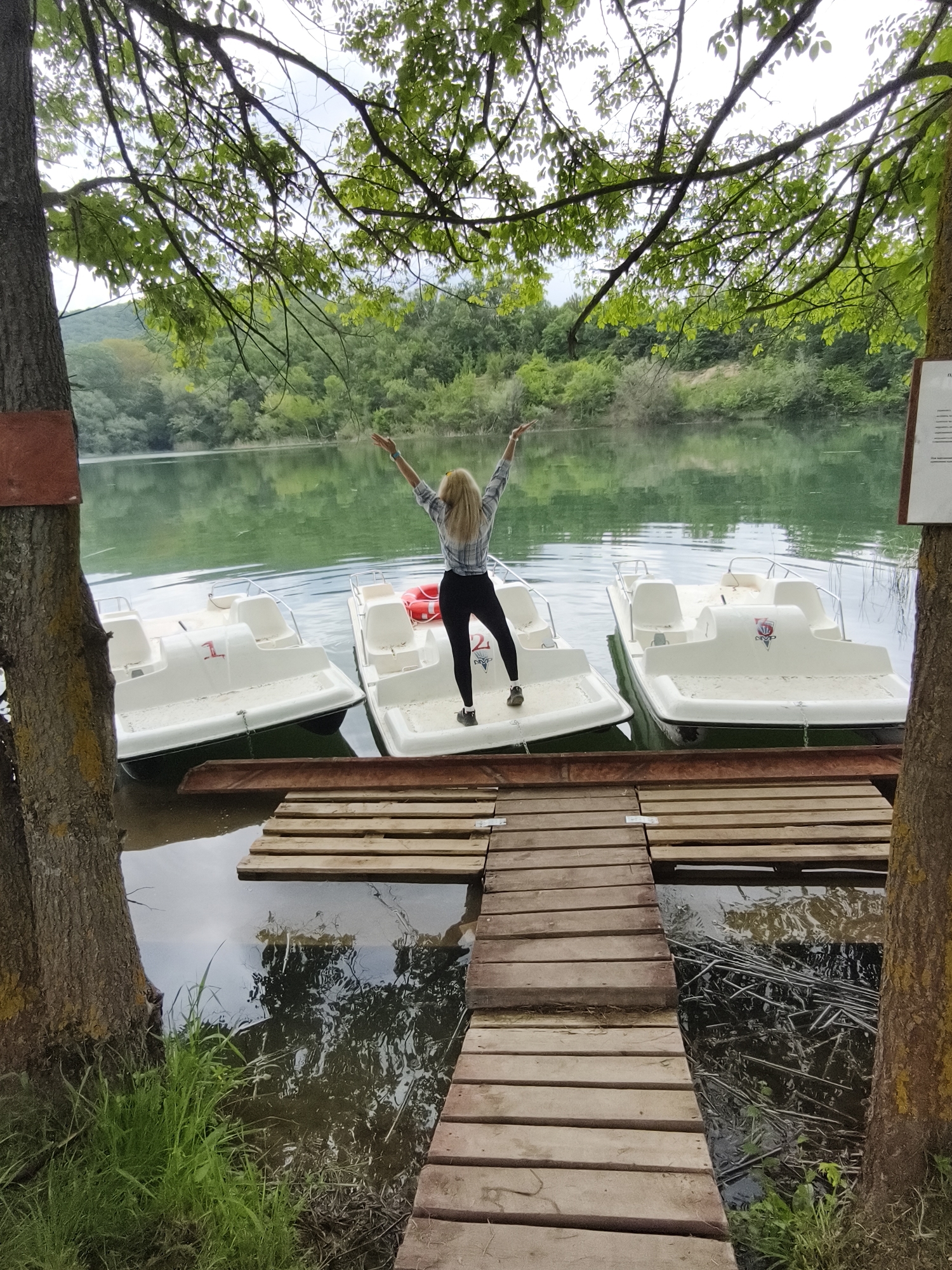 A woman stands on a white pedal boat on a lake. She is wearing black pants and a plaid shirt, and her arms are raised. The pedal boat is attached to a wooden dock. The lake is surrounded by trees. The location is Терновский округ, Ukraine.