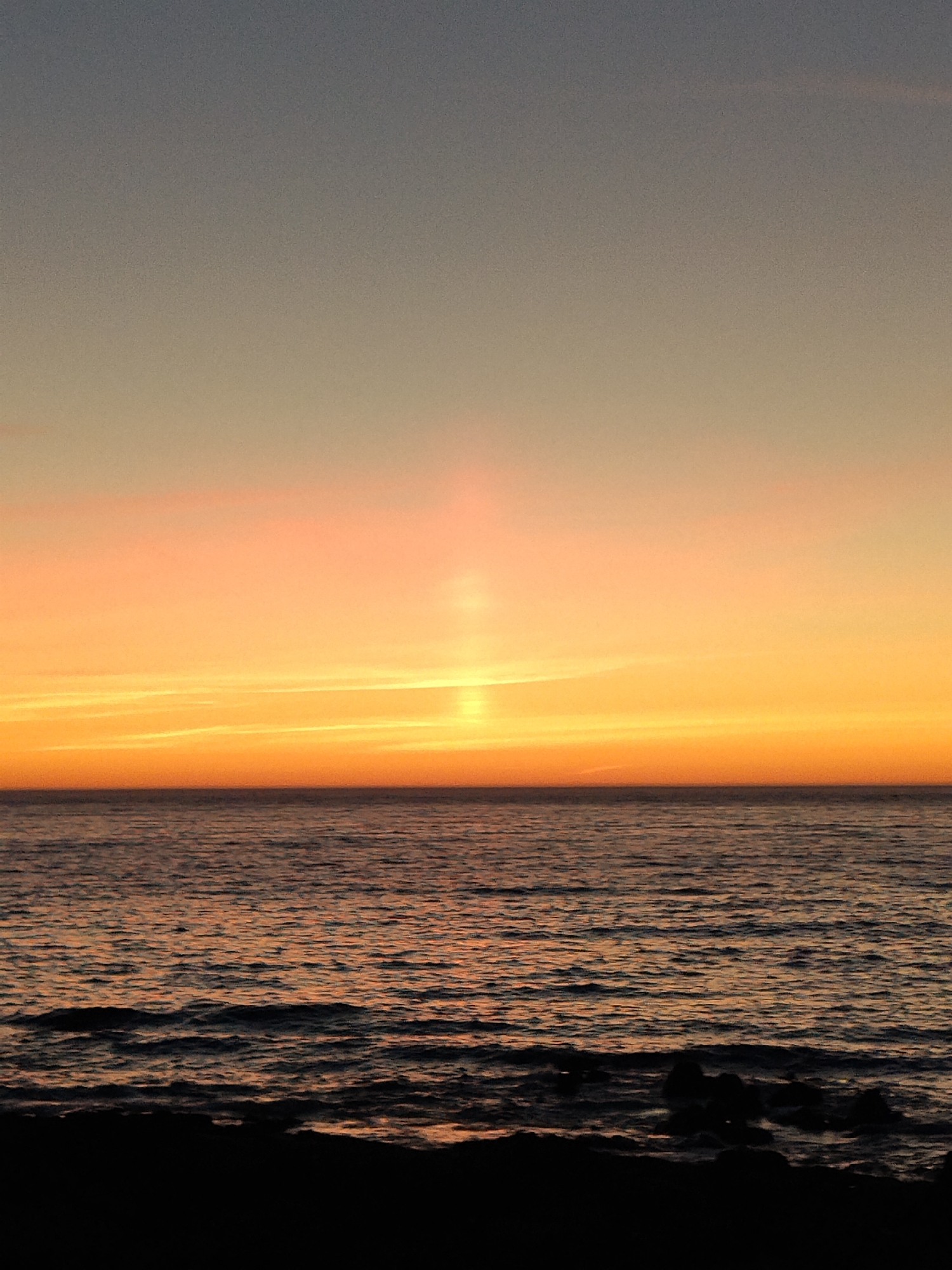 The image captures a serene sunset over the ocean. The sky transitions from a soft grey at the top to vibrant shades of orange and yellow near the horizon, with a distinct vertical beam of light emanating from the setting sun. The ocean surface is calm, with gentle ripples reflecting the warm colors of the sky. In the foreground, a dark, silhouetted shoreline is visible, with a few dark rocks peeking out of the water. The scene appears to be a natural coastal landscape, potentially a beach or rocky outcrop, characteristic of the coastline near Baiona, Spain, known for its scenic sunsets. No people or specific man-made structures are identifiable in the image.