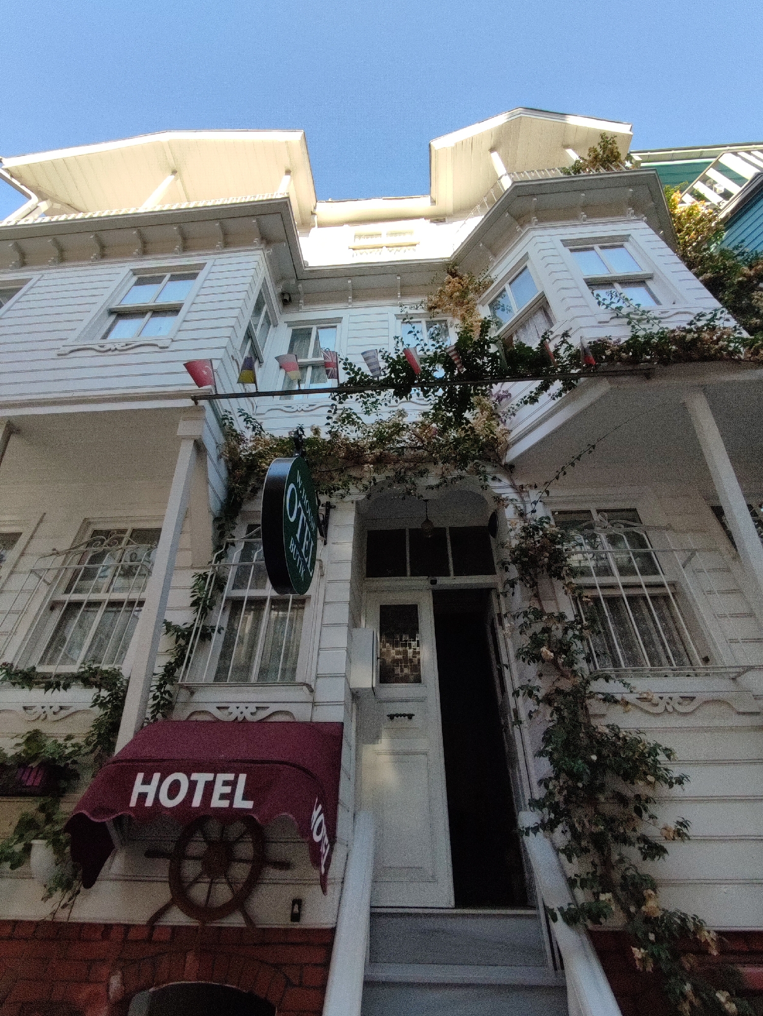 The image shows the exterior of a white, multi-story building in Kadıköy, Turkey, a coastal city on the Asian side of Istanbul. The building appears to be a boutique hotel, as indicated by a round, green sign that reads "OTEL BUTİK" and a burgundy awning above the entrance with "HOTEL" in white lettering. Small flags are strung along the upper floors, and ivy or a similar vine is growing up the sides of the building, with some flowering. The perspective is from a low angle, looking upwards towards the sky. The building has traditional architecture with prominent window frames and decorative trim. The entrance is visible with a white door and a set of stairs leading up to it.