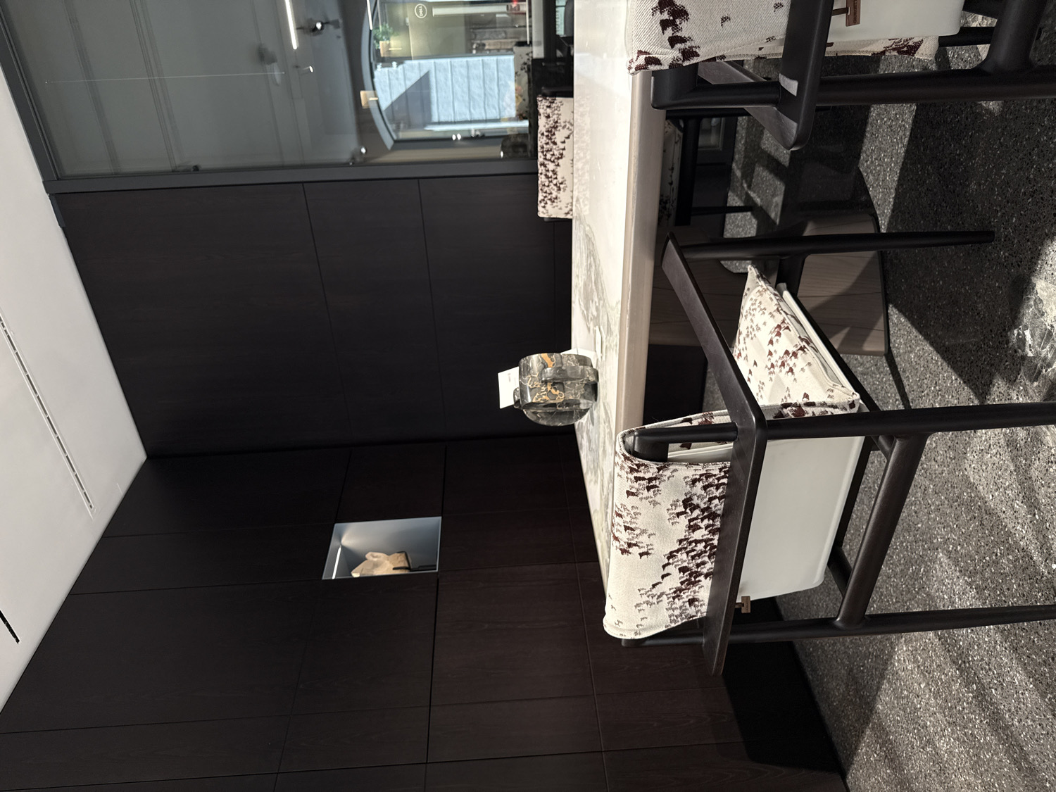 This image captures a modern and elegantly designed interior space, likely a dining area, showroom, or a high-end commercial setting in Milan, Italy. The central focus is a long, rectangular dining table with a light-colored, possibly marble or stone, top that exhibits subtle veining.
Surrounding the table are several contemporary dining chairs, characterized by their dark, possibly stained wood or metal frames with clean, angular lines. The chairs feature upholstered seats and backrests covered in a light-toned fabric with a distinctive, abstract pattern in dark brown, resembling sparse brushstrokes or small organic shapes. Some chairs also incorporate a solid white panel on the seat or back for contrast.
On the table, a decorative, clear glass or crystal object, possibly a small vase or candle holder, is visible, alongside a tiny white card with illegible text.
The background wall is clad in sleek, dark wooden paneling, creating a rich and sophisticated backdrop. Embedded within this paneling is a square, illuminated niche showcasing a light-colored, abstract sculptural object or artifact. Above the wooden panels, a large window or glass partition reflects an exterior urban scene with modern buildings and a circular sign containing an 'i' symbol, though any text is unreadable. A white ceiling segment with a ventilation grate is visible in the upper left. The floor is a dark, speckled material, possibly terrazzo or a textured carpet, which complements the overall dark and light palette of the room.
No people are present, and no specific activity is taking place, suggesting the space is either unoccupied or set up for display. The scene evokes a mood of quiet sophistication and contemporary luxury.