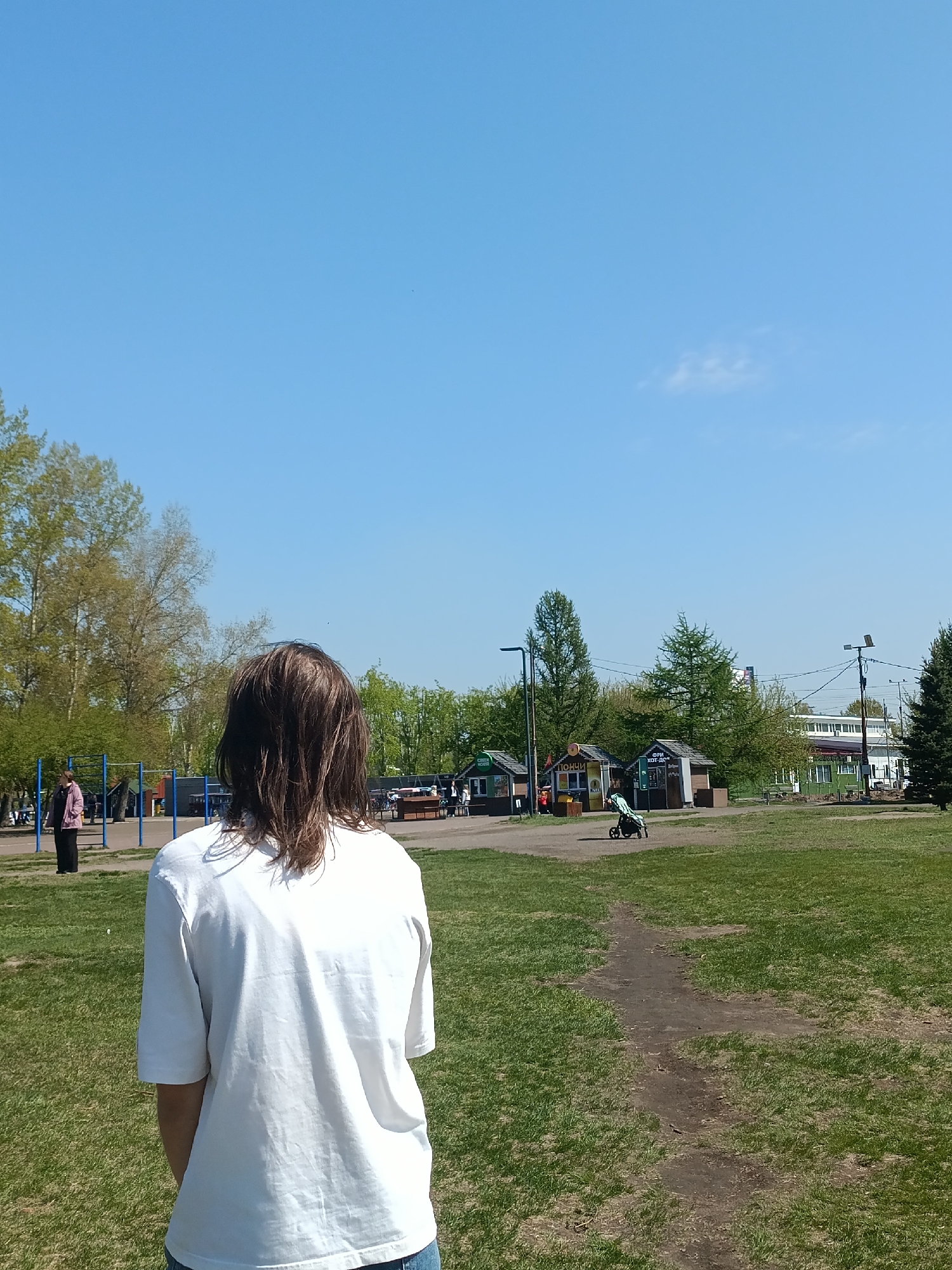 A person stands in a grassy park under a clear blue sky in Krasnoyarsk, Russia. The individual, facing away from the viewer, wears a white t-shirt and blue jeans and has shoulder-length brown hair. In the background, several small wooden kiosks line a pathway, a stroller is parked nearby. Behind the kiosks are trees and what appears to be a building in the distance. To the left, a playground structure with metal bars is visible with a person standing nearby. A dirt path leads from the foreground towards the kiosks.