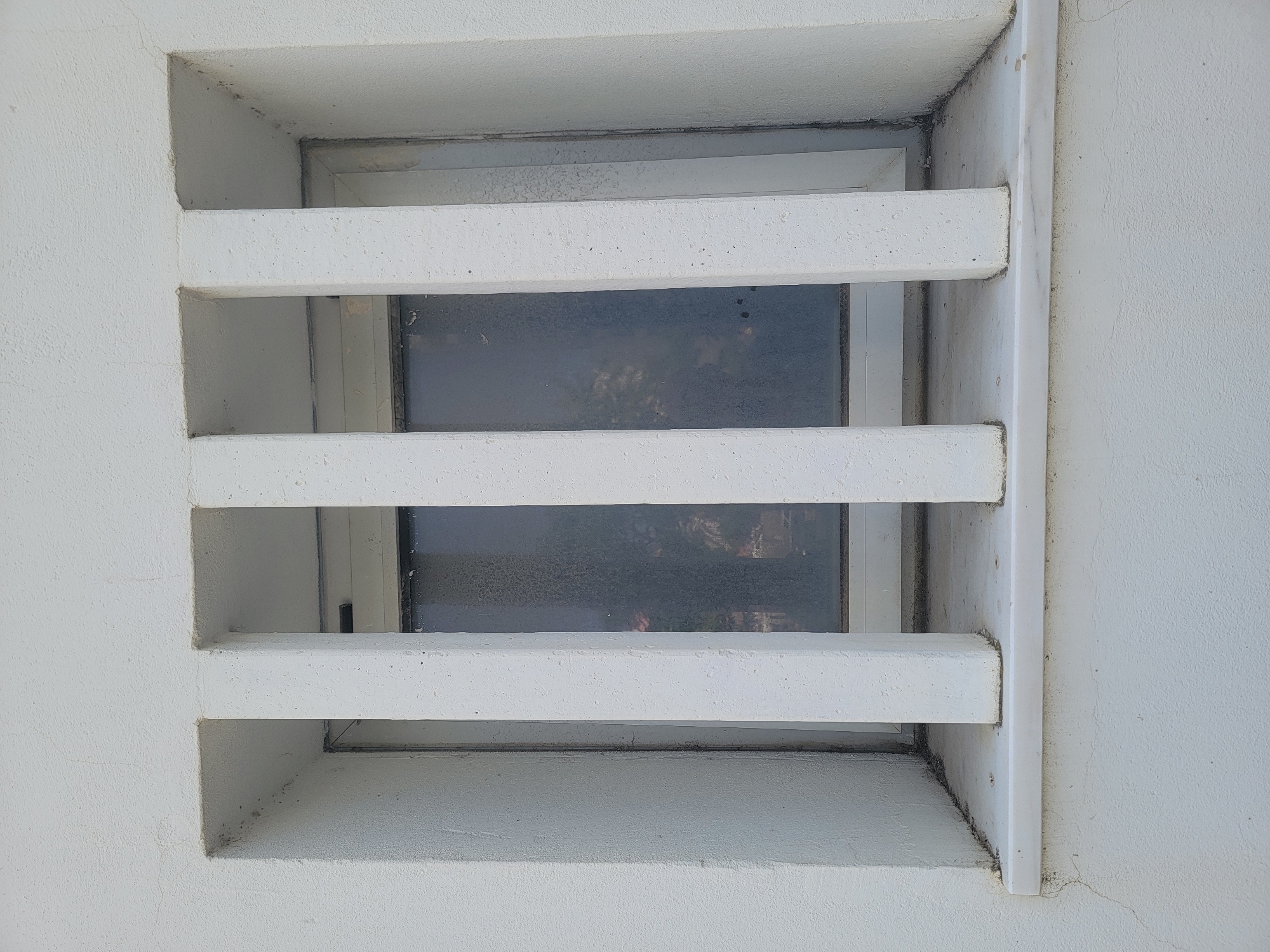 This is a close-up, eye-level shot of a small, recessed window with horizontal white louvers. The window is set into a white, textured stucco wall. The louvers are thick and appear to be made of concrete or a similar material. Through the louvers, a dark, grimy windowpane is visible, reflecting a dimly lit interior or exterior scene that is indistinct. The glass appears frosted or smudged, obscuring any clear view of what lies beyond. The setting suggests an outdoor location, possibly on a building facade. The lighting is somewhat flat, indicating it could be daytime, but the overall impression is one of a somewhat neglected or aged structure. There is no visible text in the image. The focus is on the architectural detail of the window and the surrounding wall.