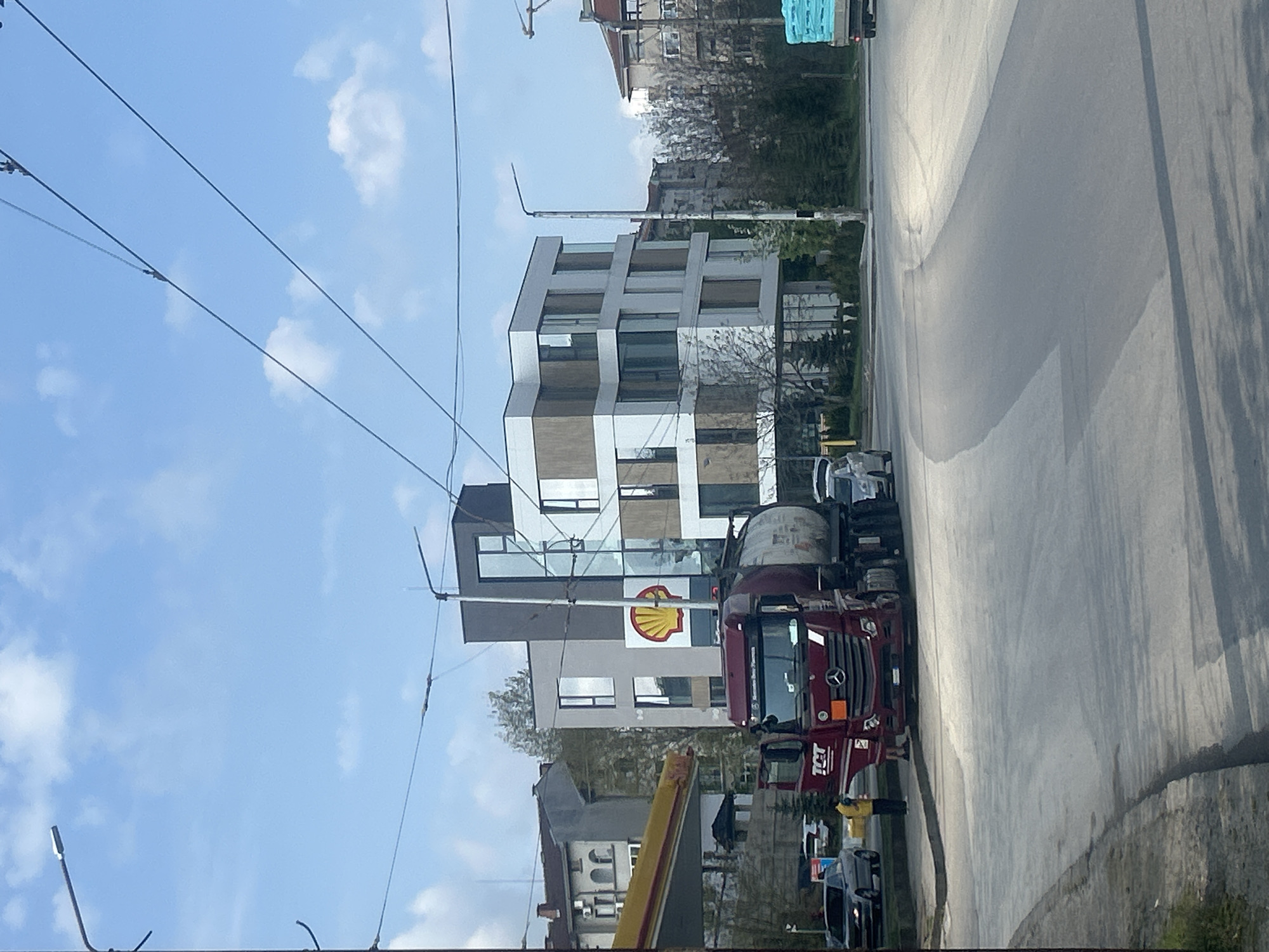 The image shows a street scene in Pleven, Bulgaria. A red Mercedes-Benz truck with a Shell logo on its tanker is parked at a Shell gas station. A modern building with a geometric facade is visible in the background. Overhead power lines are present. The sky is blue with some clouds. Other vehicles are also parked in the vicinity of the gas station. The road is paved and appears to be wet.