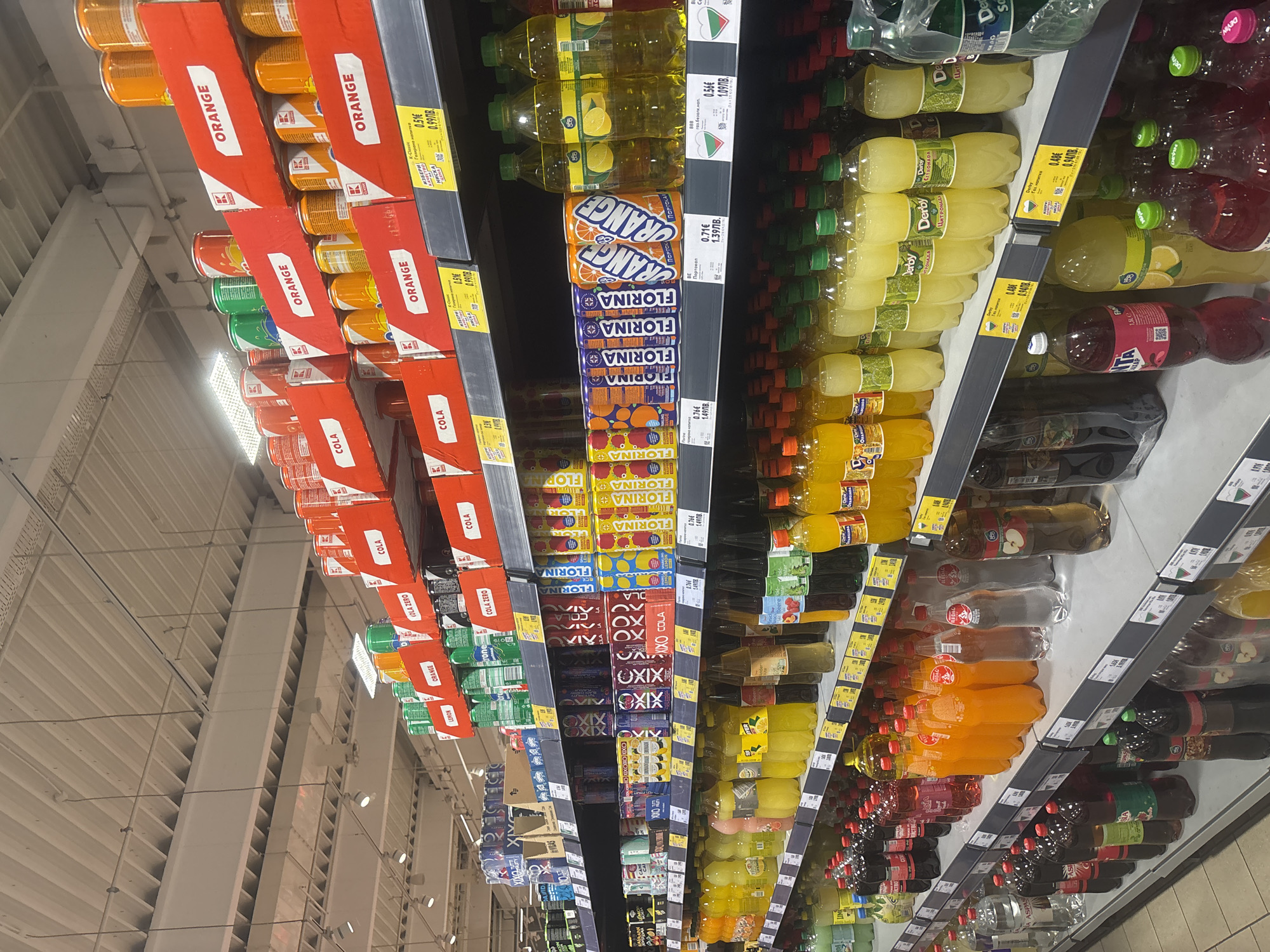 The image shows a section of a supermarket aisle in Plovdiv, Bulgaria. Shelves are stocked with various beverages, primarily soft drinks. On the left, canned drinks are arranged in multipacks, including brands like "ORANGE," "COLA," and "COLA ZERO". The middle shelves feature bottled drinks with labels indicating "FLORINA" and "ORANGE". Bottles of "Derby" lemonade in different flavors line the adjacent shelf. Price tags in Bulgarian Lev (ЛВ) and Euro (€) are visible beside the products, offering cost comparisons. The lighting is bright, typical of a retail environment. The angle suggests the photograph was taken looking down the aisle from one end. No people are visible in the shot.