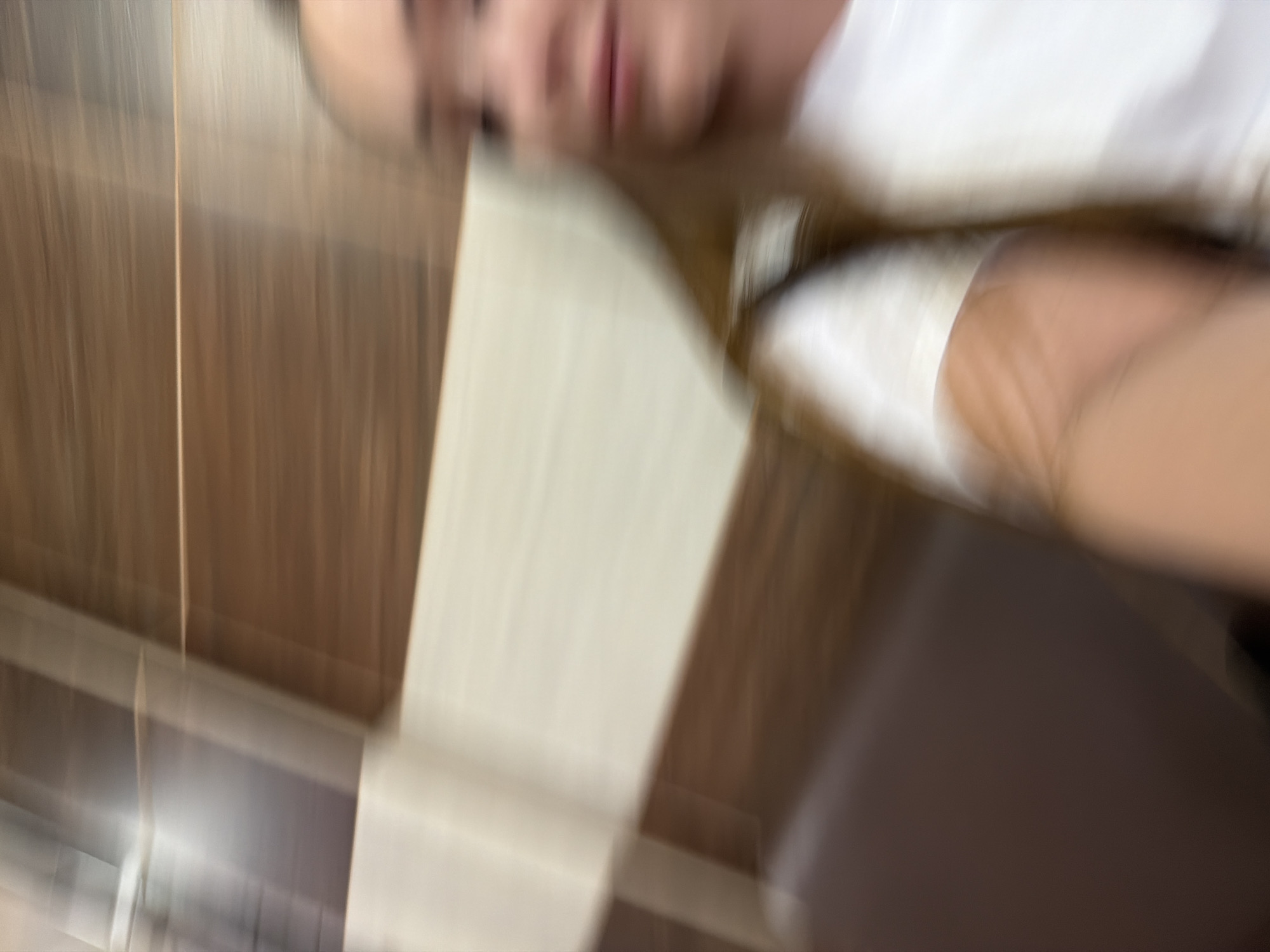 The image is blurred, but it shows a person wearing a white shirt. A wall with wood paneling is partially visible. The location of the image is unknown and cannot be identified with reasonable certainty.