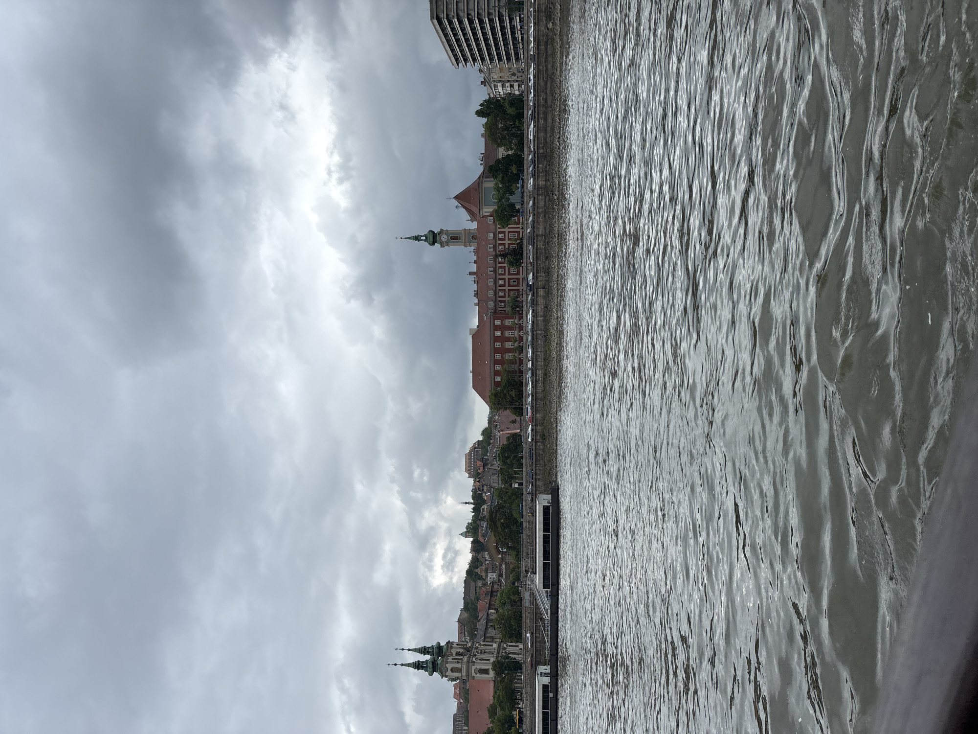 The photo captures a view of the city of Budapest, Hungary, from the Danube River. Overcast skies create a somber atmosphere. Buildings line the riverbank, including distinctive structures with towers and spires. The water surface reflects the sky's gray tones. In the middle distance, detailed architectural elements are visible on buildings of varying heights. Closer to the viewer, the river's texture is characterized by ripples and movement.
