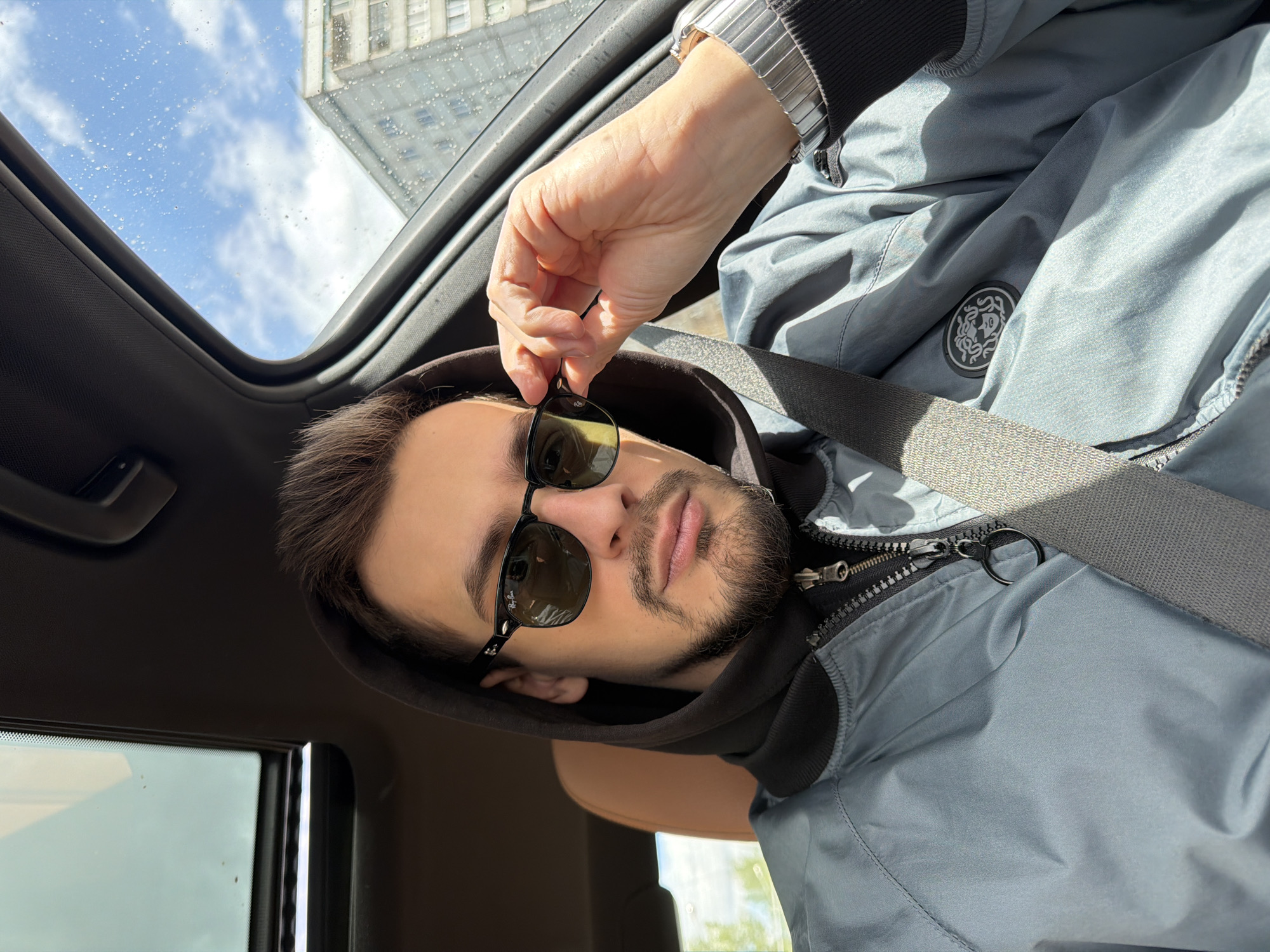 A man in a car is putting on sunglasses. He is wearing a blue-grey hooded jacket and a seatbelt. His left hand is holding the sunglasses. Through the open sunroof, a building and blue sky with white clouds are visible. Based on the architectural style of the building visible through the sunroof, this image was not taken in the city of Krasnoyarsk, Russia. The architectural style of the building suggests that this photo was taken in a North American city.