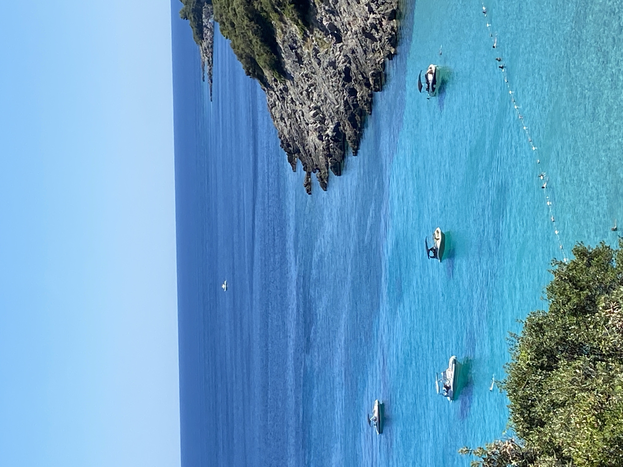 In the city of Dubraljevina, Montenegro, the scene unfolds with a panoramic view of a coastal landscape on a clear, sunny day. The upper portion of the image is dominated by a vast expanse of brilliant blue sky, transitioning into the deep blue of the open sea.
Closer to the shore, the water becomes remarkably clear and turquoise, revealing the sandy bottom in places. Several small boats are dotted across this vibrant water. One white boat with a black canopy is anchored in the foreground, its reflection shimmering on the surface. Another similar boat is positioned slightly further out, and two more are visible in the distance.
Along the right side of the frame, a rocky coastline curves into the sea, covered in lush green vegetation. The rocks appear weathered and uneven. A thin rope with buoys stretches across the water in the middle ground, marking a boundary. Within this roped-off area, several people are swimming and enjoying the water. They are small in the frame, scattered and engaged in leisure activities. In the far distance, a lone white boat sails on the horizon. The foreground is partially obscured by the dense green foliage of a tree.