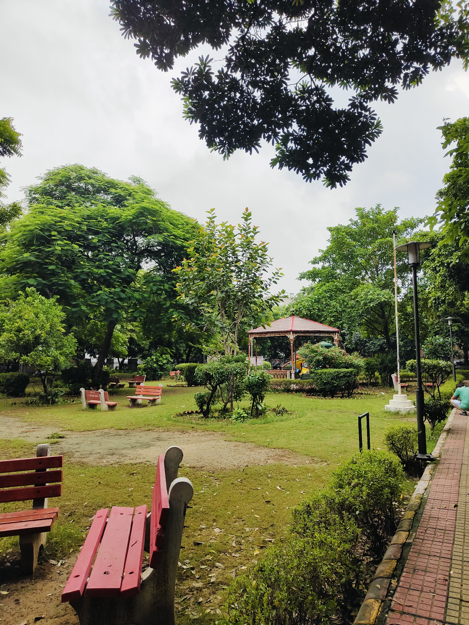 The image depicts a park scene in Gurgaon, India, with a focus on a covered pavilion and seating areas. In the foreground, two red park benches with concrete bases are visible. One bench is angled towards the viewer from the left, and another is in the center, facing away from the viewer. The ground is covered with patchy green grass and a dirt path. In the mid-ground, a large, multi-tiered red-roofed pavilion with intricate white latticework stands prominently. Several other red park benches are scattered throughout this area. Surrounding the pavilion and benches are manicured green bushes and lush trees. A lamppost with a black metal pole and a light fixture is positioned to the right of the pavilion. Further to the right, a paved walkway made of red tiles with a yellow border runs alongside a row of green bushes. A person wearing a green t-shirt and blue shorts is crouched down near the end of the walkway, facing away from the camera. The sky is overcast with a pale grey hue. The overall impression is of a well-maintained public park during the daytime.