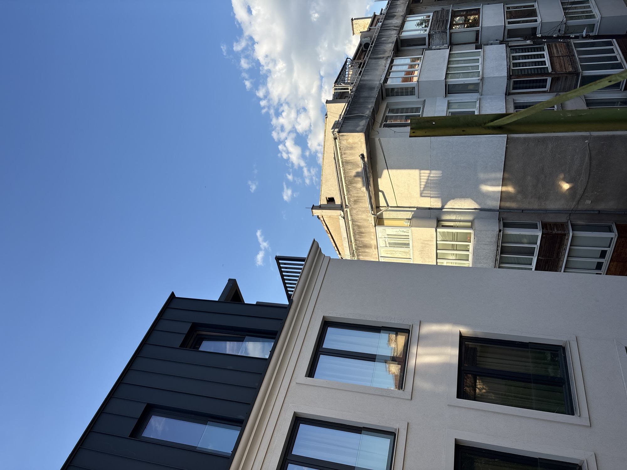 A low-angle shot shows a juxtaposition of three buildings against a bright blue sky with scattered clouds, likely in Sofia, Bulgaria, given the architectural style. In the foreground, the lower section of a modern building is visible. It features a dark gray, possibly metal, exterior with horizontal panels and windows reflecting the sky. Adjacent to it is a renovated building with a white textured façade, also featuring modern black-framed windows. Above these, an older building with a worn, concrete exterior is positioned, showing a variety of windows, some with external shutters and other modifications. A green pipe runs along the façade. The composition emphasizes the contrast between modern renovation and older urban architecture.