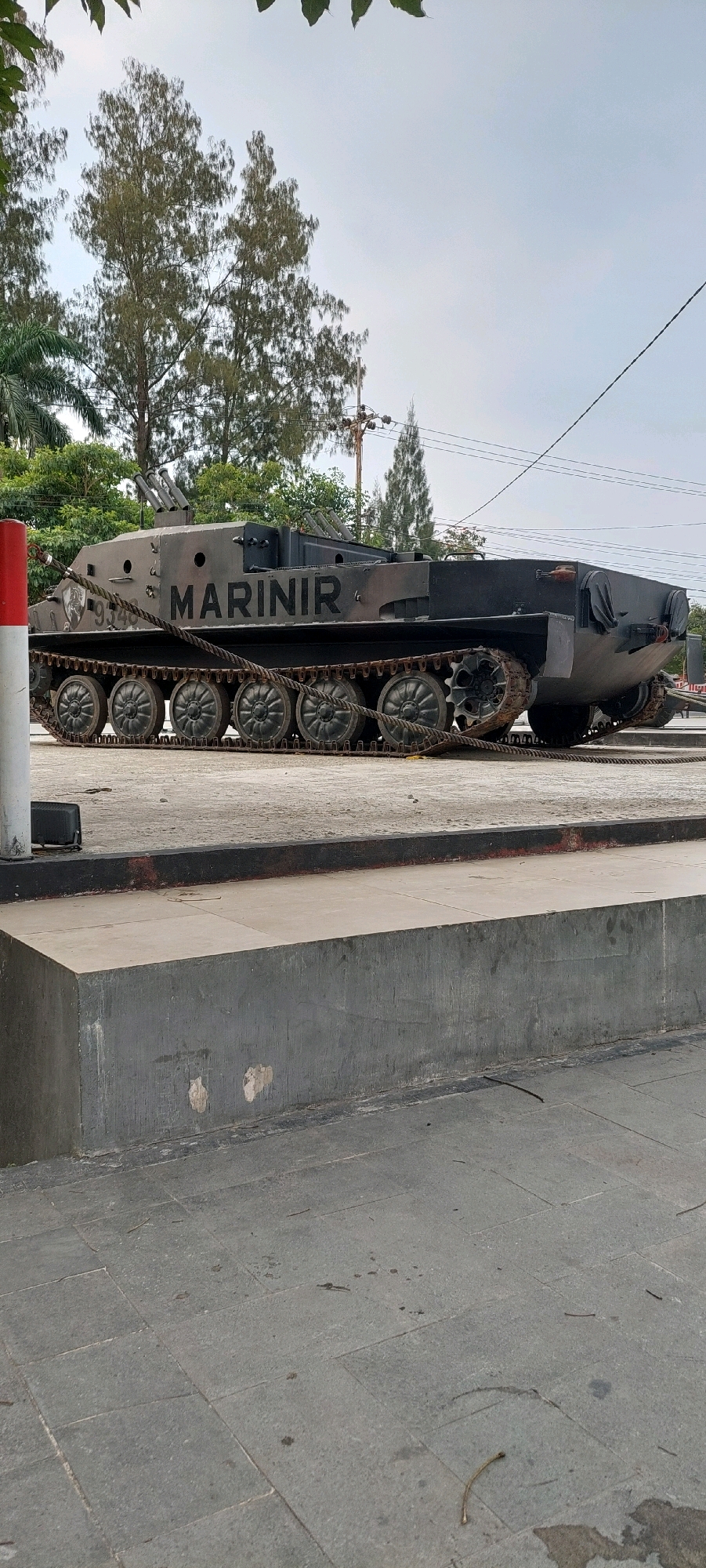 A static display of an amphibious assault vehicle, painted in a camouflage pattern of dark grey and black, is situated outdoors on a paved surface. The vehicle bears the inscription "MARINIR" in large, black capital letters on its side. The track system of the vehicle is visible, with multiple road wheels and a heavy-duty track.
In the foreground, a low, dark grey concrete structure runs horizontally across the image, with a lighter grey paved surface extending further into the foreground. A red and white striped pole stands vertically on the left side of the frame, near the front of the vehicle, and is connected to the vehicle by a thick rope.
Behind the vehicle, a line of trees with slender trunks and feathery foliage forms a backdrop. Electrical poles and wires are also visible against the overcast sky. The ground is a mixture of concrete slabs and gravel, with some fallen leaves and debris scattered about. The overall scene suggests a military display or monument in a public outdoor space. The location is specified as Unknown, Unknown.