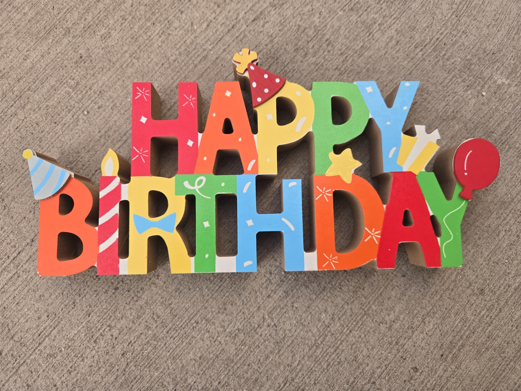 A colorful "Happy Birthday" sign is placed on a textured gray surface. The sign is made of wooden letters, each decorated with various patterns and colors. Some letters feature additional embellishments like party hats, a candle, a bow tie, a star, and a balloon.