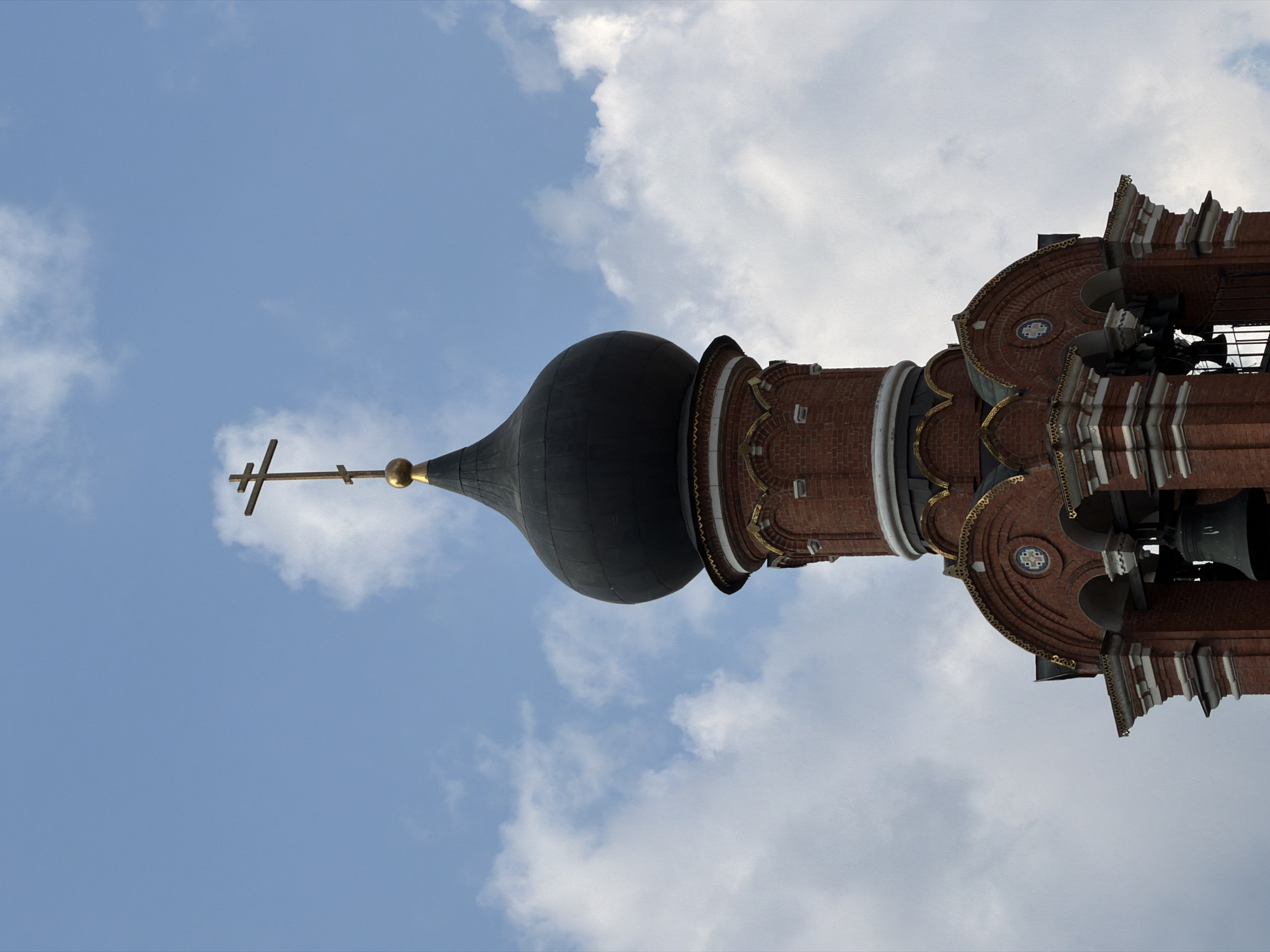 The image shows a close-up of the bell tower of the Church of the Assumption of the Blessed Virgin Mary in Khimki, Russia. The tower is made of red brick and has a black dome topped with a golden Orthodox cross. The sky is blue with scattered white clouds. The bell tower has multiple levels with arched openings where bells are visible. Decorative elements, including gold trim, are present on the tower. The overall scene is peaceful and depicts a religious structure against a cloudy sky.