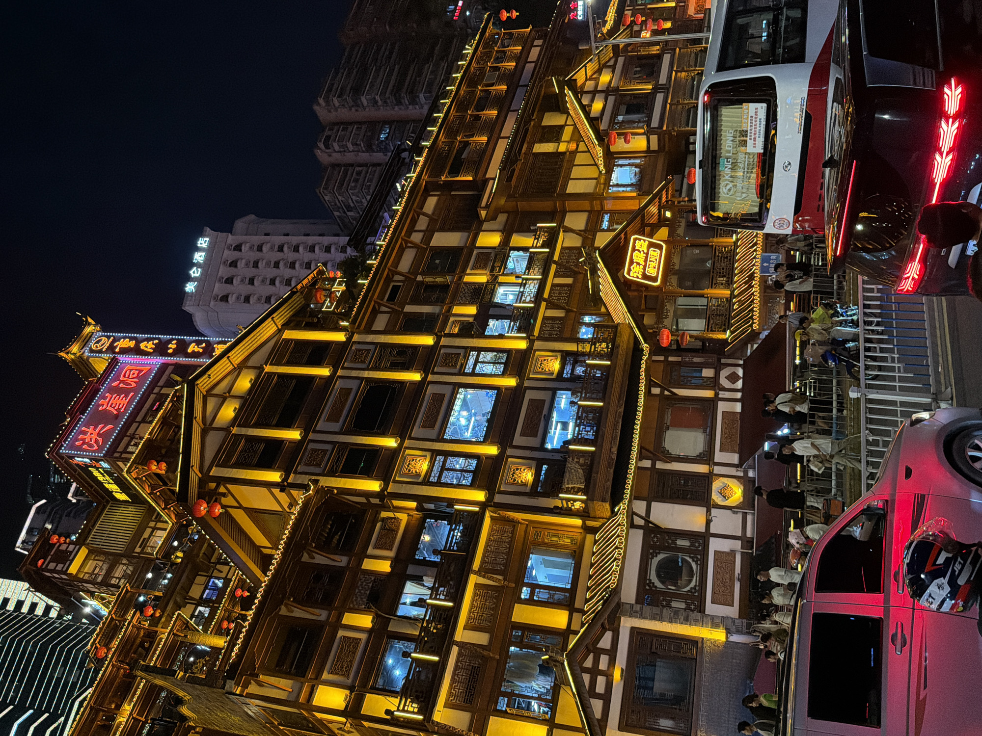 This media file depicts a night scene in Yuzhong District, China, specifically showcasing the vibrant nightlife and architecture of Hongyadong. The image is dominated by a multi-story building with traditional Chinese architectural elements, illuminated by numerous warm yellow lights that highlight its intricate wooden facade and windows. Red lanterns are strategically placed, adding to the festive ambiance.
In the foreground, a bright red bus with "KING LONG" branding is visible, indicating public transportation. Beside it, a pink van with red tail lights is parked. Several people are seen near the bus and the van, some appearing to be passengers or onlookers, suggesting a lively gathering or transit point.
Several signs with Chinese characters are displayed on the buildings, including "洪崖洞" (Hongyadong), "重庆小天" (Chongqing Xiao Tian), "美仑酒店" (Meilun Hotel), "徐鼎盛" (Xu Dingsheng), and "渝味川菜" (Yuwei Sichuan Cuisine). These signs suggest that the area is a popular destination for dining and accommodation, with Hongyadong being a prominent landmark. The "C位江装" sign might refer to a specific establishment or promotion. The overall atmosphere is one of bustling urban activity at night, characterized by bright lights, distinctive architecture, and the presence of both vehicles and pedestrians. The weather appears clear, allowing for visibility of the illuminated cityscape.