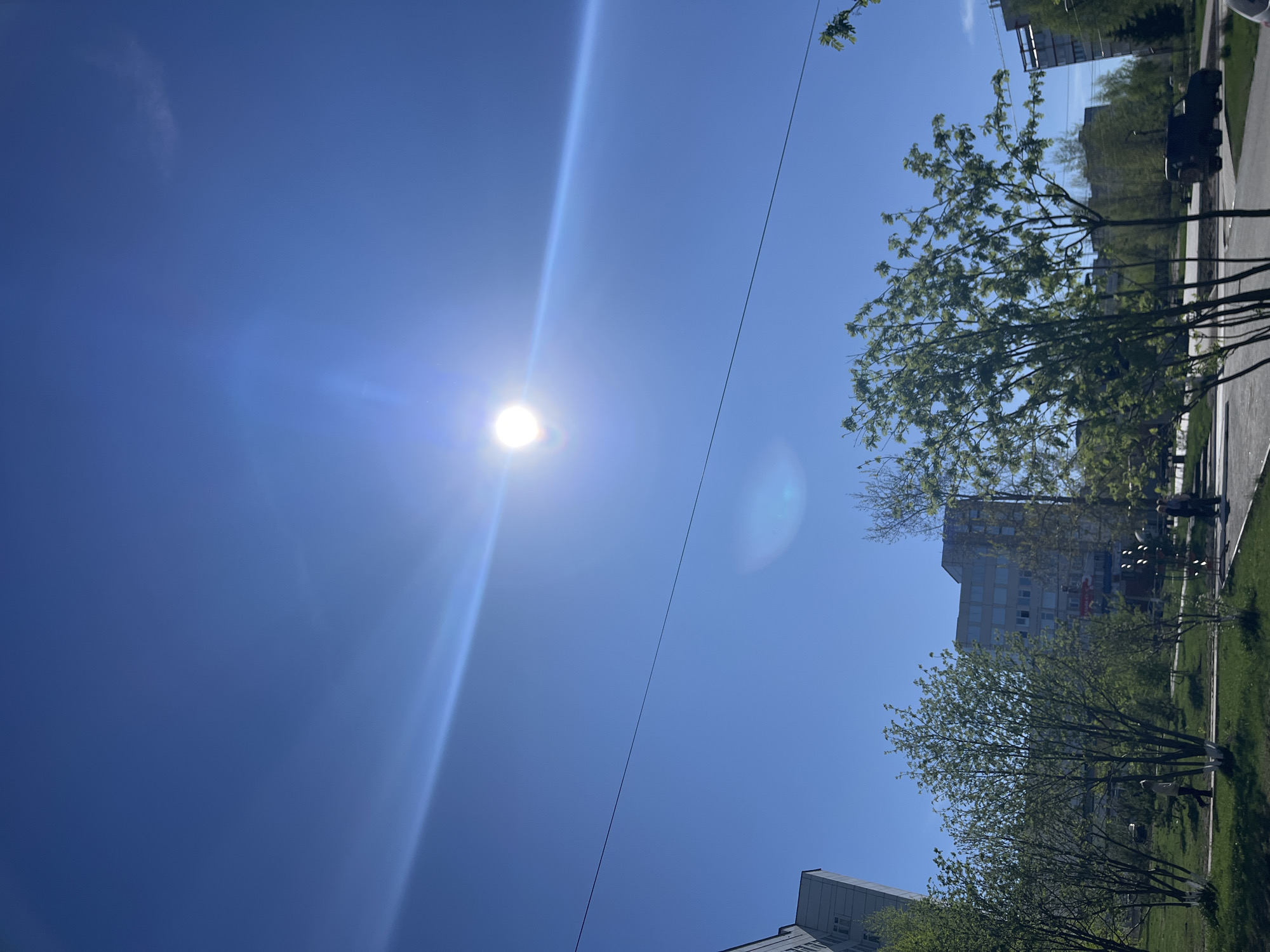 The image shows a street scene in the city of Chaikovsky, Russia on a bright, sunny day. The camera is pointed upwards at the sky, capturing the sun with a lens flare and a clear blue sky above. A power line runs diagonally across the sky. Trees line the street to the right, partially obscuring a tall, gray apartment building with multiple windows. People can be seen walking along the sidewalk. The image is oriented vertically.