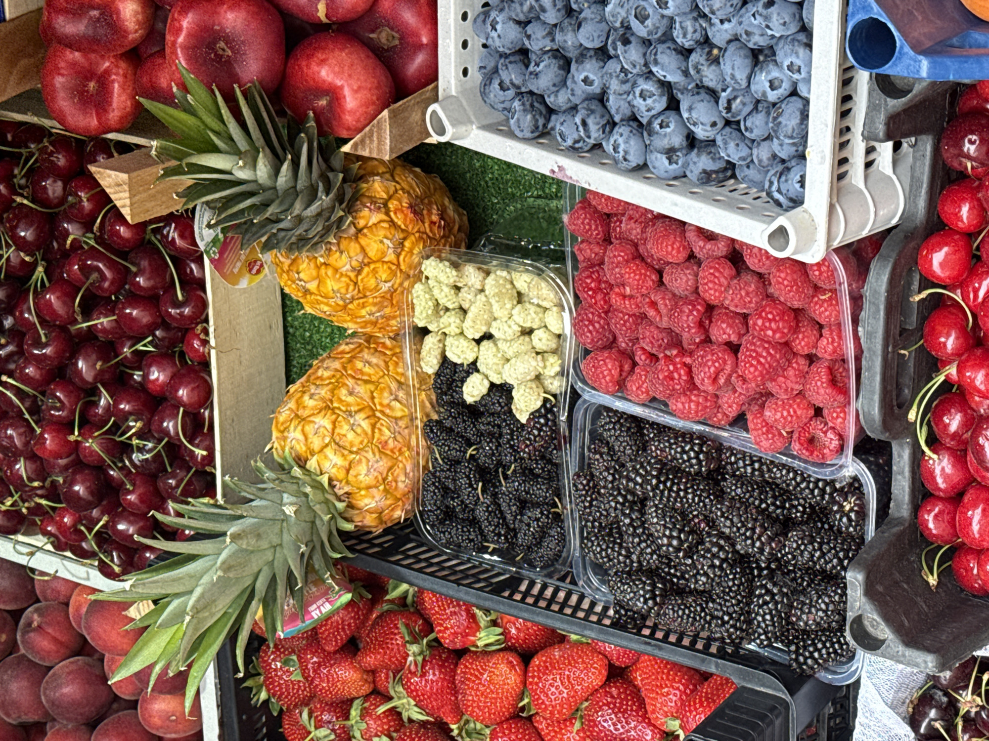 This is a close-up, eye-level shot of a fruit stall in Saint Petersburg, Russia, displaying an abundant assortment of fresh produce. The scene is dominated by various berries, pineapples, and what appear to be peaches or nectarines. In the foreground and mid-ground, several black plastic crates hold generous portions of bright red strawberries, plump blackberries, vibrant red raspberries, and a container of white mulberries. To the right, a white plastic crate is filled with a large quantity of blue blueberries. Two ripe pineapples are positioned in the upper half of the frame, their spiky green crowns contrasting with their textured yellow skins. In the background, larger fruits are visible, including a pile of deep red cherries and round, reddish-pink fruits, possibly peaches or nectarines, in wooden crates. The stall appears to be situated outdoors, possibly at a market, with a green surface, likely artificial turf or fabric, serving as a base for some of the produce. There are no people visible in the image, and the focus is entirely on the colorful display of fruits.