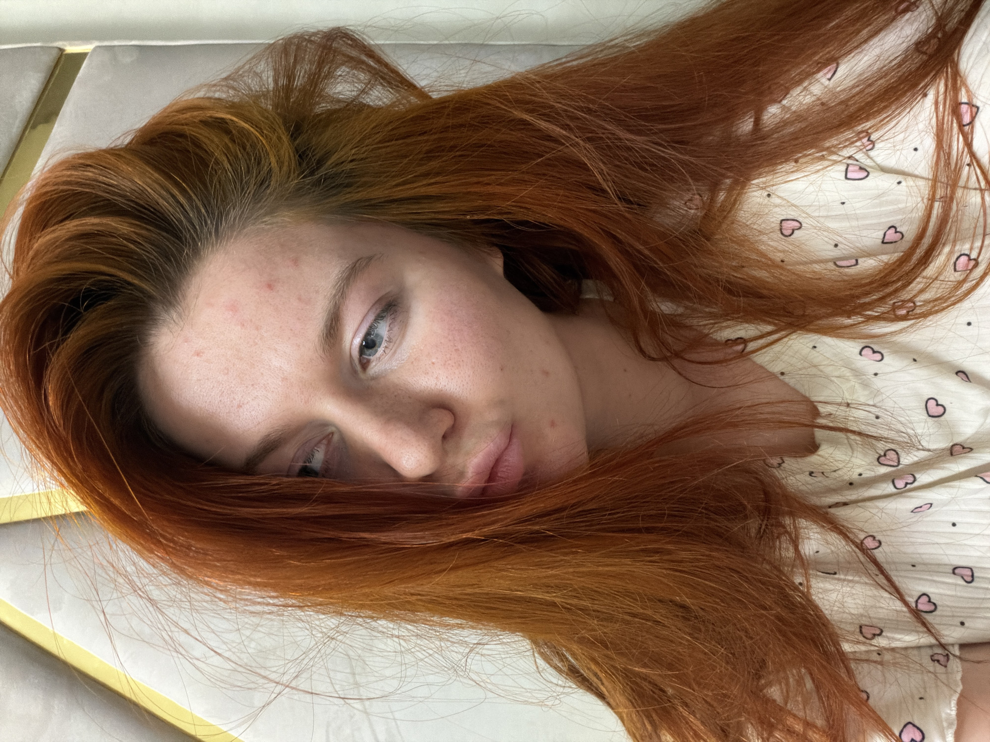 A close-up, overhead shot shows a young woman with long, bright red hair. Her hair is tousled and falls across her face and shoulders, partially obscuring her features. She is looking slightly upwards and to her left, with her lips pursed in a pout. Her skin appears fair, with some visible freckles and a few small blemishes on her forehead and cheeks. She is wearing a light-colored garment with a pattern of small pink hearts and black dots. The background is softly lit, with a textured surface that appears to be a wall or headboard, possibly with a golden trim. The lighting suggests an indoor setting, likely a bedroom. There is no visible indication of the city of Krasnodar, Russia, or any other specific geographical location.