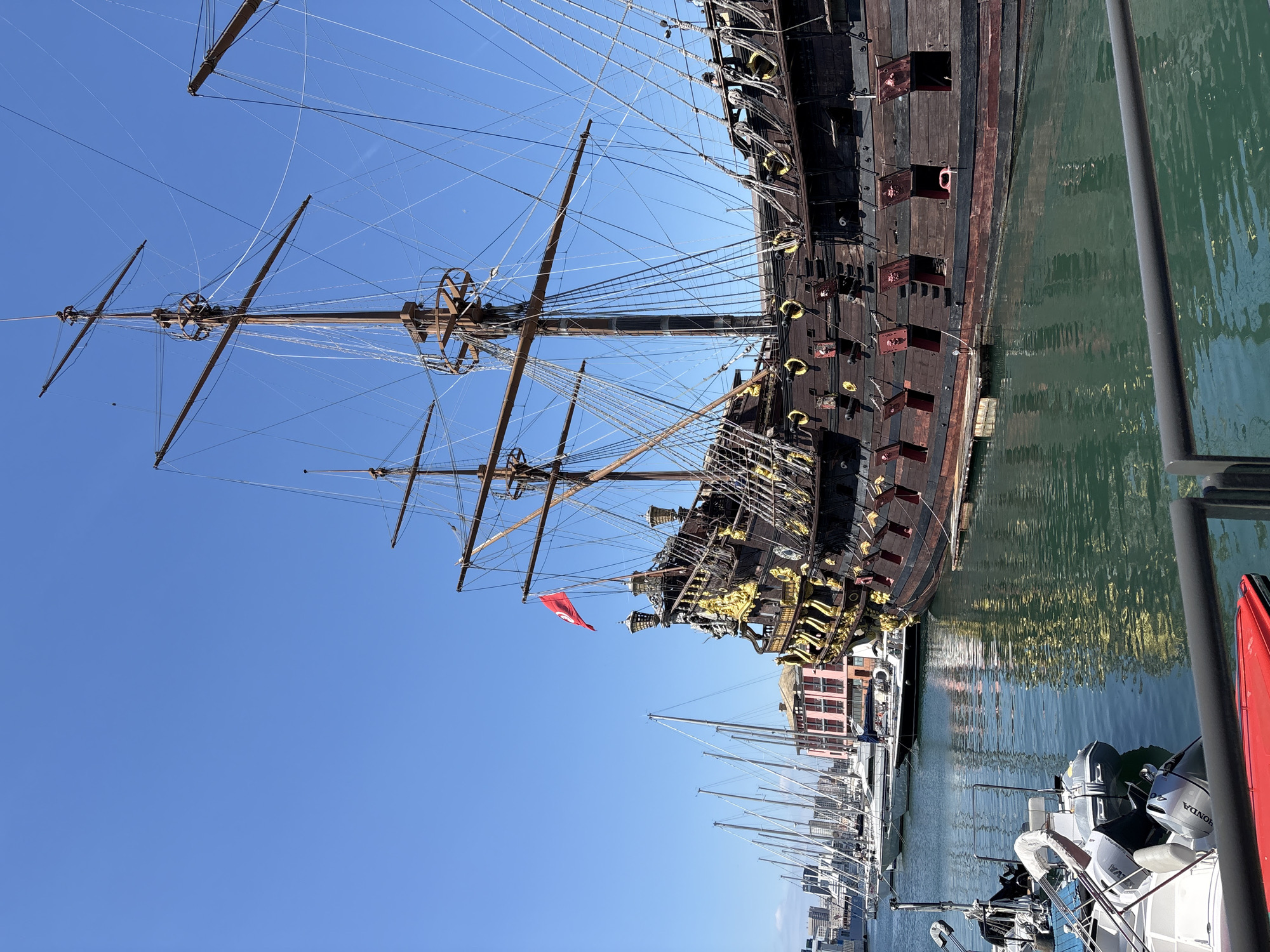 The image shows a full-scale replica of a 17th-century galleon docked in the harbor of Genoa, Italy. The ship is a dark wood color with gold decorative elements. It features multiple masts and intricate rigging. A red flag is visible at the ship's stern. The galleon is docked alongside a row of modern sailboats and powerboats. In the background, there are buildings on the waterfront. The sky is clear and blue. The water in the harbor reflects the sunlight.