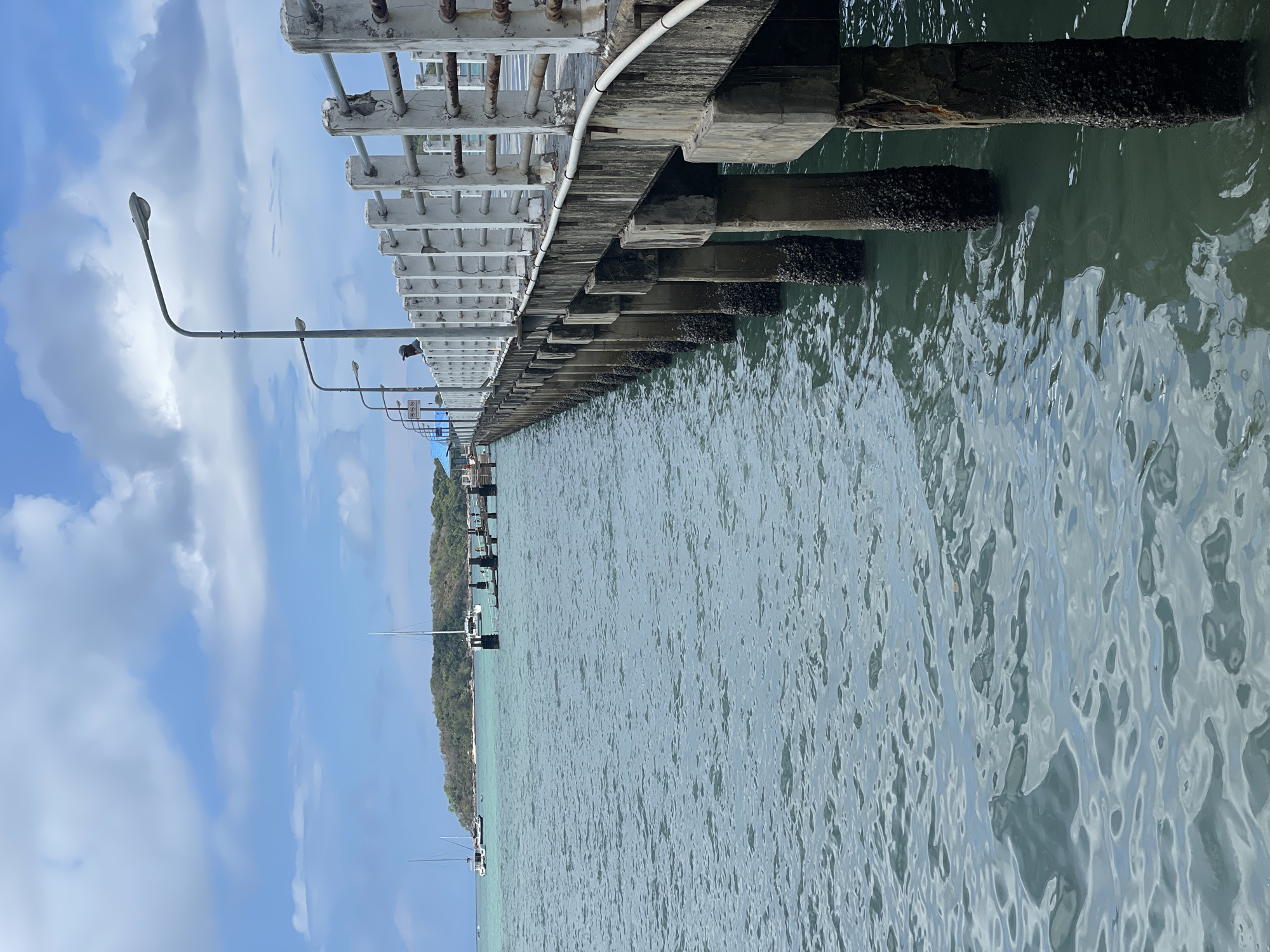 A long pier extends into the water, lined with streetlights and a white, lattice-like structure overhead. The supports of the pier are visible, some encrusted with marine growth. Beyond the pier, there's a coastline with trees and some docked boats. The water shows a rippled surface, reflecting light. The sky is blue with scattered clouds. The photo was taken at the Rawai Pier in Thailand.