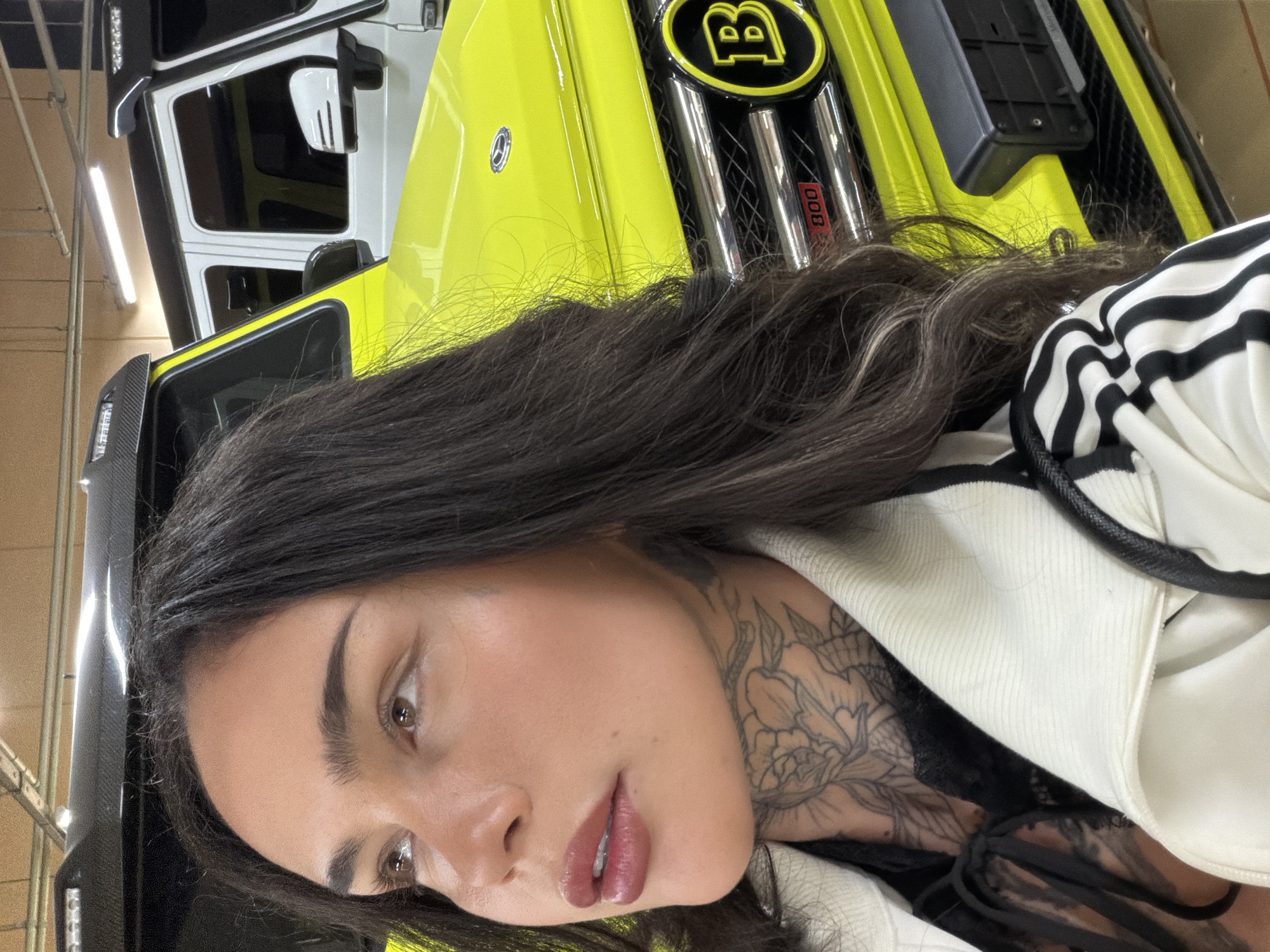 A medium shot captures a light-skinned person with dark hair and tattoos on their neck. They are wearing a white jacket with black stripes. Behind them is a bright yellow car, identified by a "B" logo as a Brabus vehicle, likely a customized Mercedes-Benz G-Wagon. The location is undetermined, with the absence of any information to confirm Seoul, South Korea.