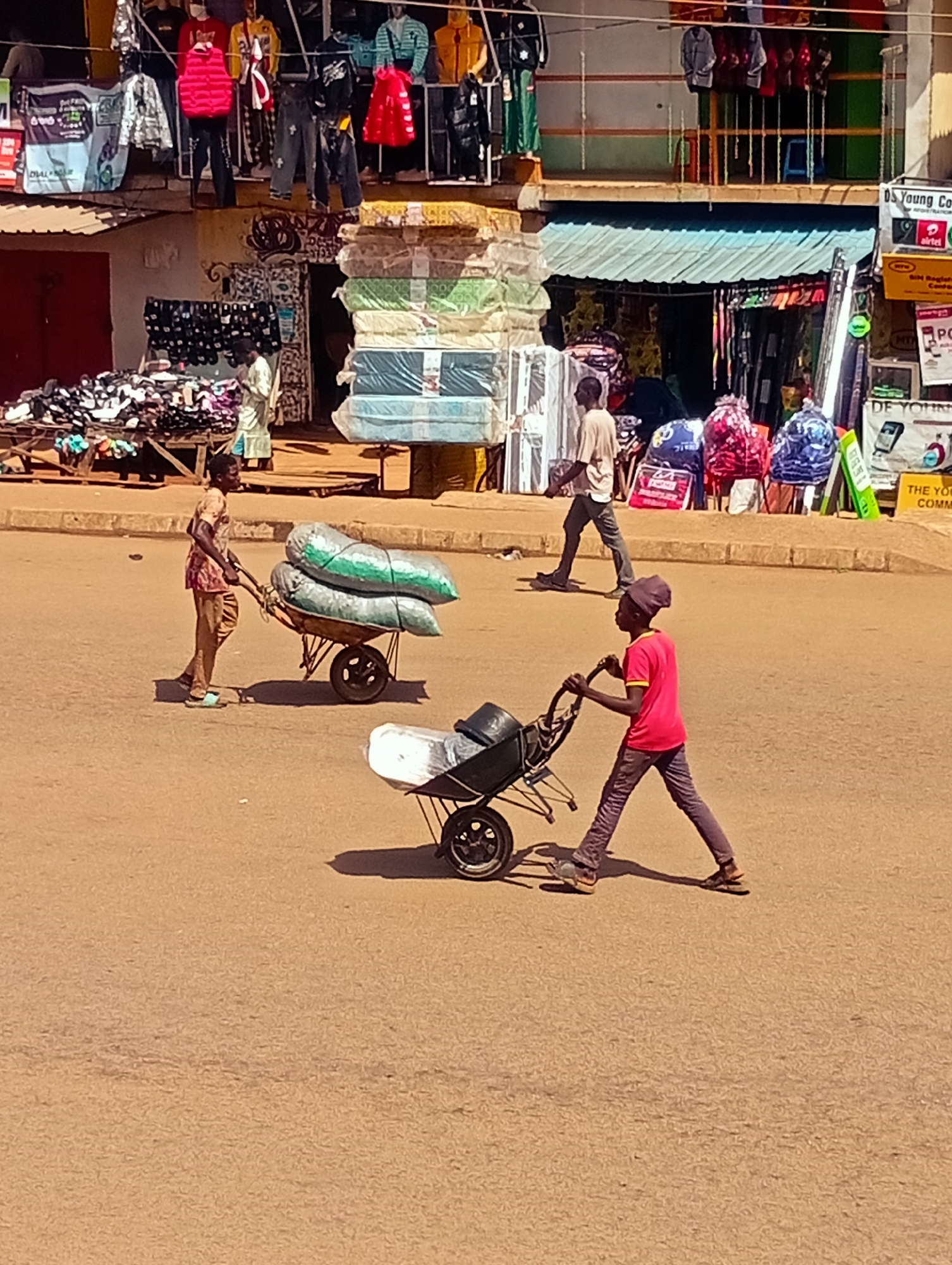 In the city of Bukuru, Nigeria, a busy street scene unfolds. Two young boys navigate the dusty road, each pushing a wheelbarrow. The boy on the left, clad in a patterned shirt and light-colored pants, is pulling a wheelbarrow loaded with two large sacks of what appears to be produce, possibly yams, tied with green netting. The boy on the right, wearing a bright red shirt and dark pants, is pushing an empty wheelbarrow. Behind them, stalls overflow with merchandise. To the left, a stall displays stacks of brightly colored mattresses, some wrapped in plastic. Further back, other stalls are visible, featuring clothing and other goods. The overall impression is one of vibrant commercial activity in a bustling Nigerian city.