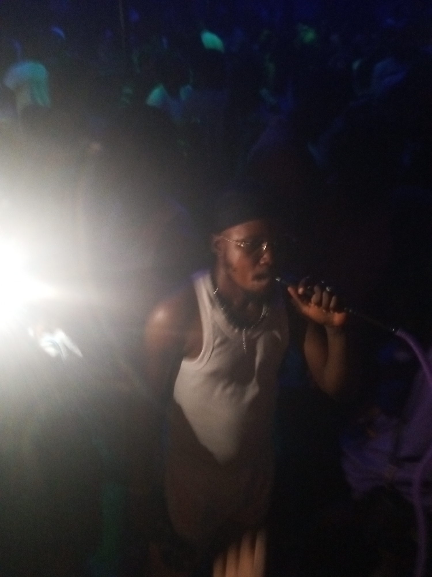 A man is singing into a microphone. He is wearing a white tank top and glasses and is in front of a crowd. The location of the image is a dark room in Lokoja, Nigeria. The crowd is barely visible in the dark.