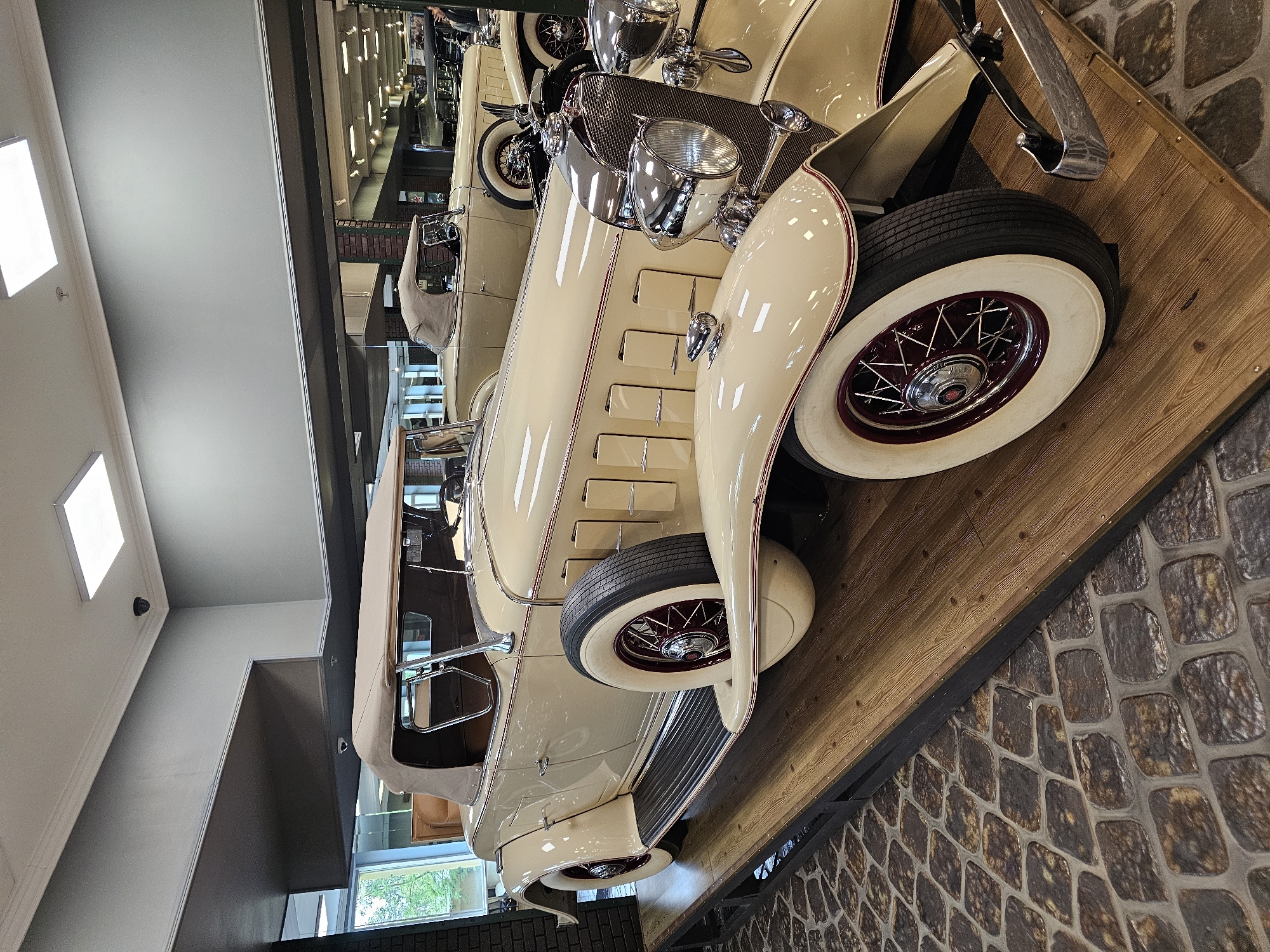 The image displays a vintage cream-colored car with red rims and white sidewall tires. The car is positioned on a wooden platform with a dark border, adjacent to a stone-like tiled area. The location appears to be indoors, with a view through a window revealing greenery outside. There are other vintage cars visible in the background, suggesting a museum or exhibition setting. The image was taken in Архангельское, Russia.