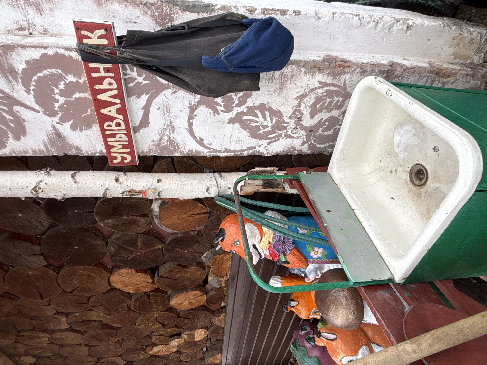 The image depicts an outdoor washbasin ("Umyvalnik" in Russian) mounted on a decorative structure, likely in the city of Chaikovsky, Russia. The washbasin itself is white with a green exterior, appearing aged. Below it, there are carved figures of animals, possibly foxes and birds, painted in bright colors. The structure is mounted on a wall made of stacked logs, a common feature in Russian rural architecture. Above the washbasin, a bag and a blue hat are hanging. A red sign with white Cyrillic letters "УМЫВАЛЬНИК" (Umyvalnik) is attached to the wall. The wall itself is decorated with painted patterns in brown and white.