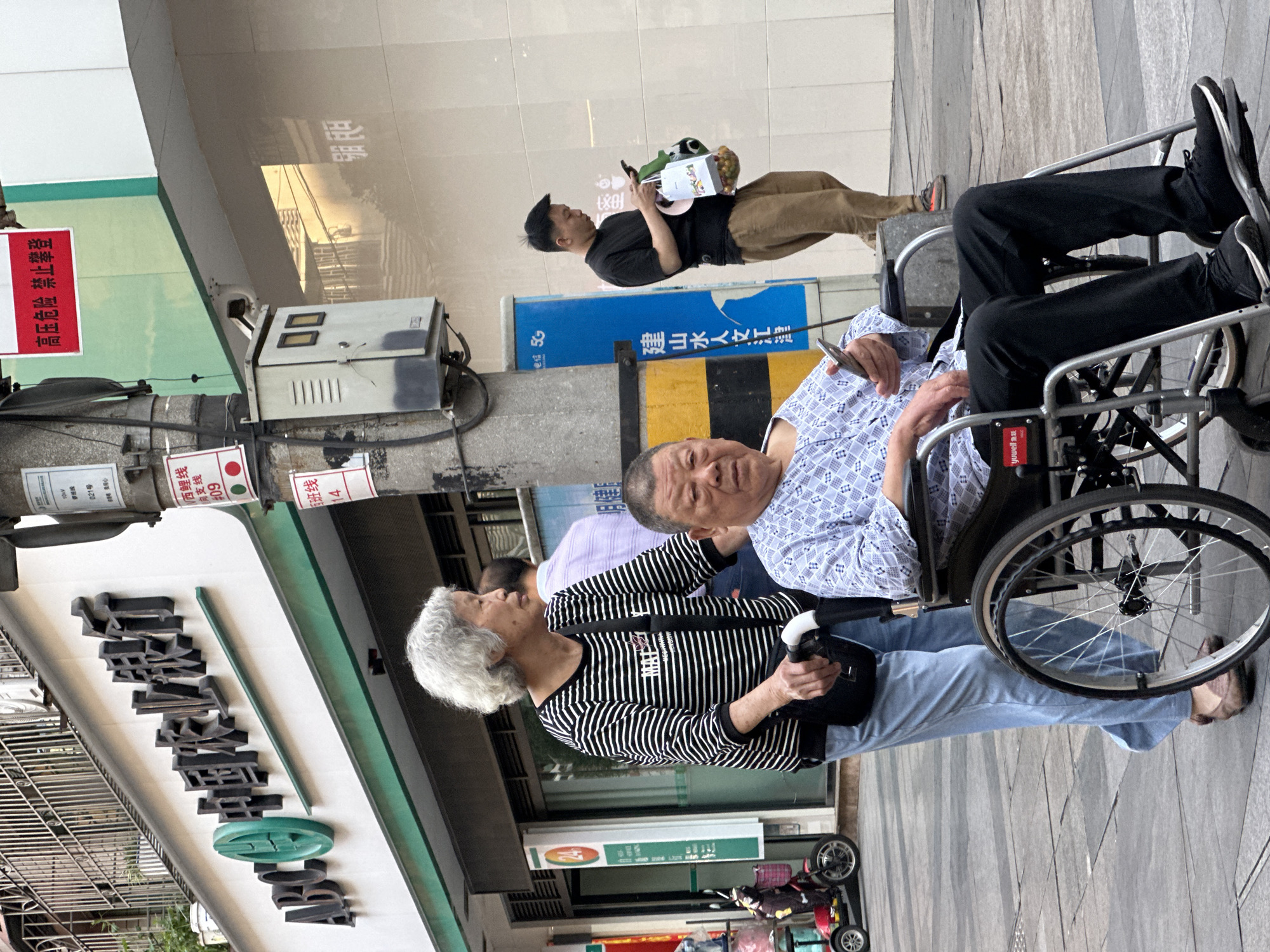 The image shows a scene on a city street, likely in Jiangjin, China, given the Chinese signage. A man in a wheelchair is the primary subject; he is wearing a light blue patterned shirt and appears to be looking at his mobile phone. The wheelchair has a person's legs resting on it; they are wearing black pants and black sneakers. An older woman in a black and white striped shirt and light blue jeans is walking next to the wheelchair. She is holding a black bag and looking in the direction of the sign. To the left, there is a storefront with Chinese characters and a green trim. A sign to the far left reads "高压危险 禁止攀登," which translates to "High voltage danger, do not climb." Another sign has the words “ADCO 中国农业银行”, “yuwell 鱼跃”, “月健”, “修班线”, “用心”, “班线”, “电信 5G”, “建山水人文汇”, and “西鲤线 向支线”, which identifies that the branch offers agricultural services. Another man is seen in the background with a black shirt and khaki pants, also looking at his mobile phone and carrying a bag with various items. The ground is paved, and a small vehicle is partially visible at the bottom of the image.