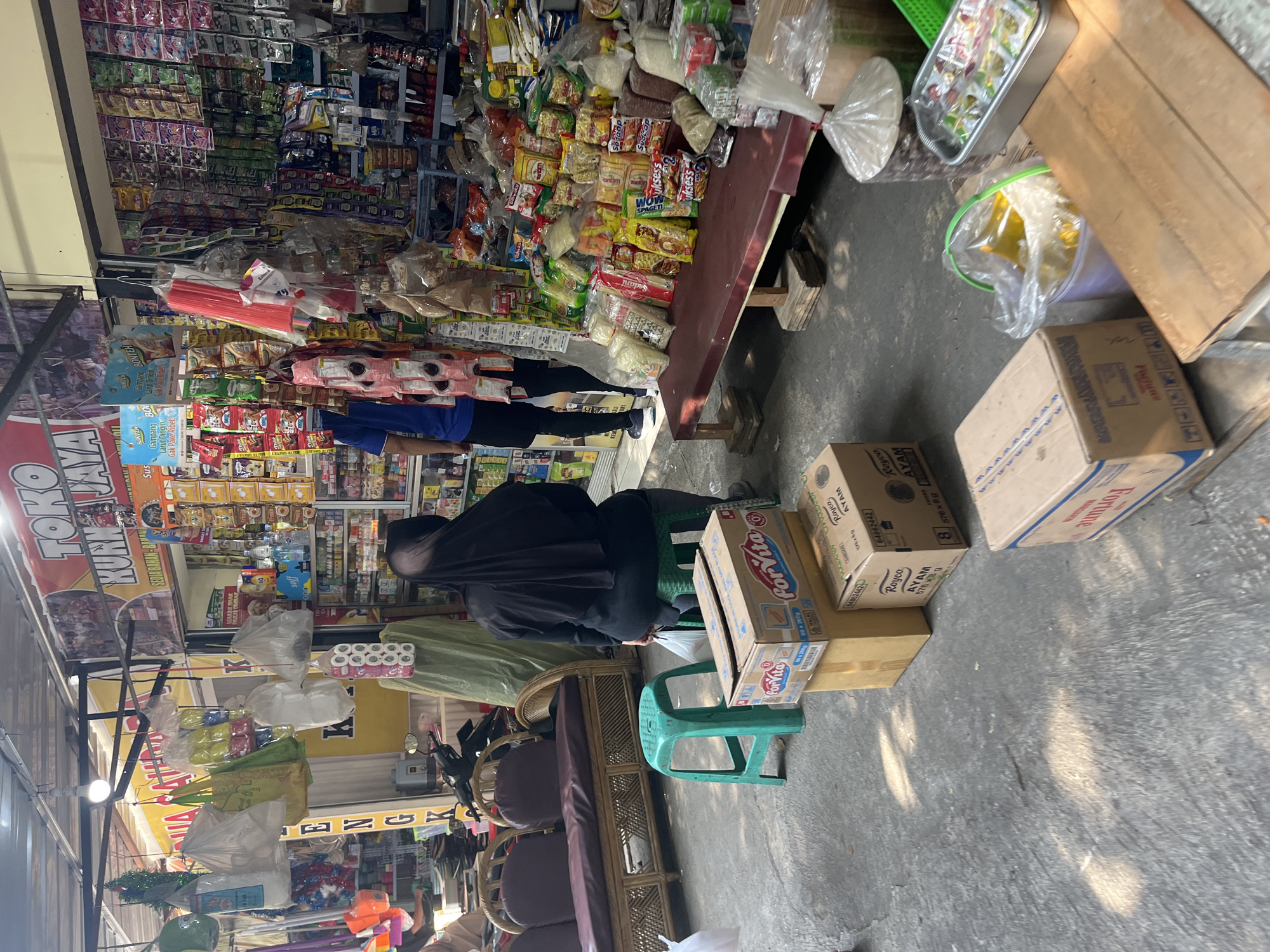 The image depicts a street-level convenience store or market stall in Bandung City, Indonesia. The stall is packed with various goods, including packaged snacks, drinks, household items, and cooking ingredients, displayed on shelves and a long wooden counter. Various plastic bags, some filled with products and others empty, hang from the ceiling. In the foreground, a woman wearing a black hijab and dark clothing is seated on a green plastic stool. Next to her, on the ground, are several cardboard boxes, one of which is labeled "ForVita" and another "Royco AYAM". A green plastic stool is positioned near the boxes. Behind the seated woman, a portion of a woven couch or bench is visible. Further back, a man wearing a blue t-shirt and dark shorts stands facing the shelves. A motorcycle is parked behind a green tarp. Sunlight casts dappled shadows on the concrete ground. The overall scene suggests a busy, informal retail environment.