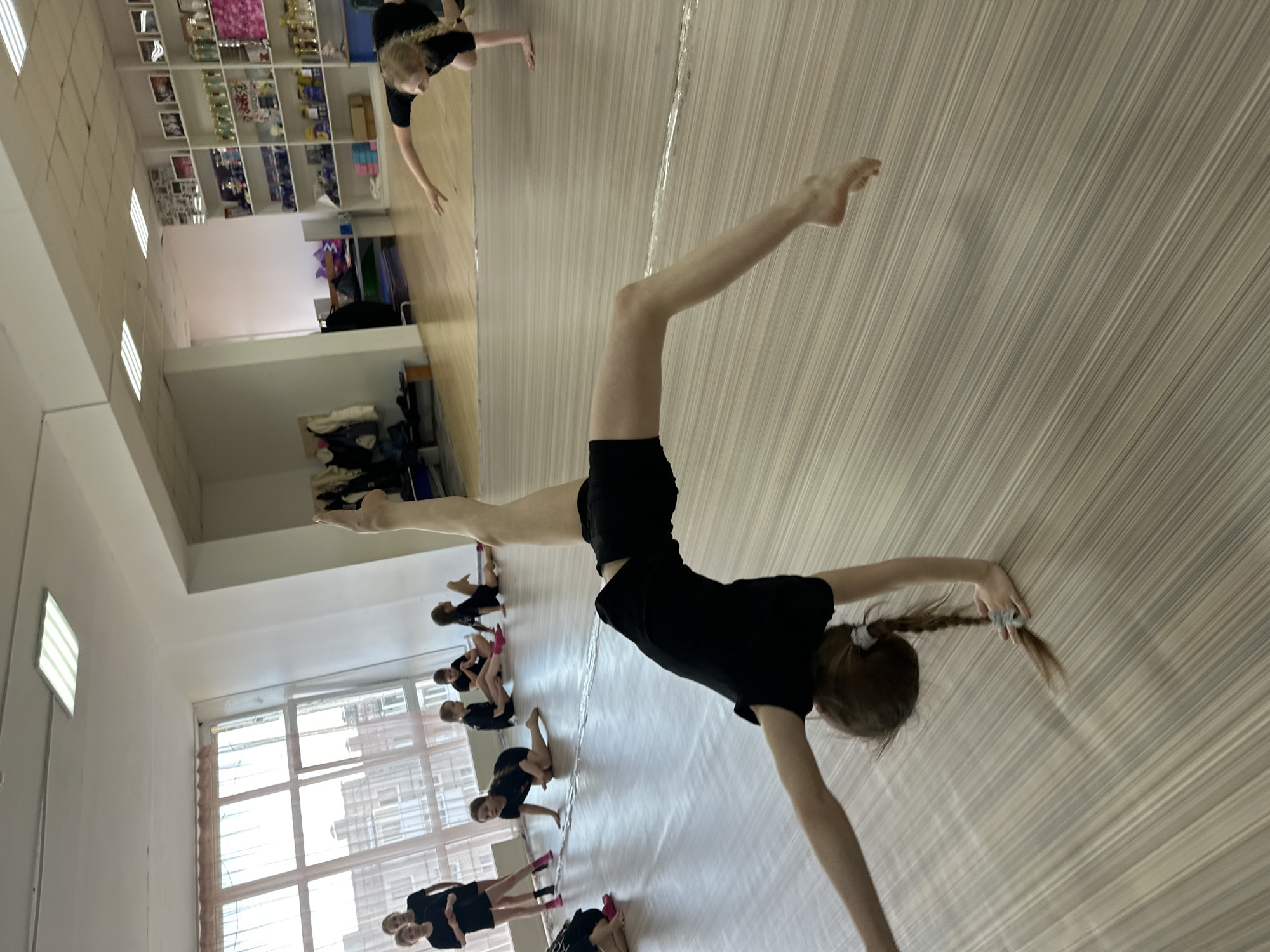In a dance studio in Votkinsk, Russia, a group of young female gymnasts are practicing. One gymnast is in the foreground, performing a handstand with one leg extended straight up and the other forward. She is wearing a black leotard and has her hair in a braid. Other gymnasts, also in black leotards, are visible in the background, practicing various poses near a window. The studio has a wood-patterned floor and a mirror on one wall, reflecting some of the gymnasts and a shelf full of trophies.