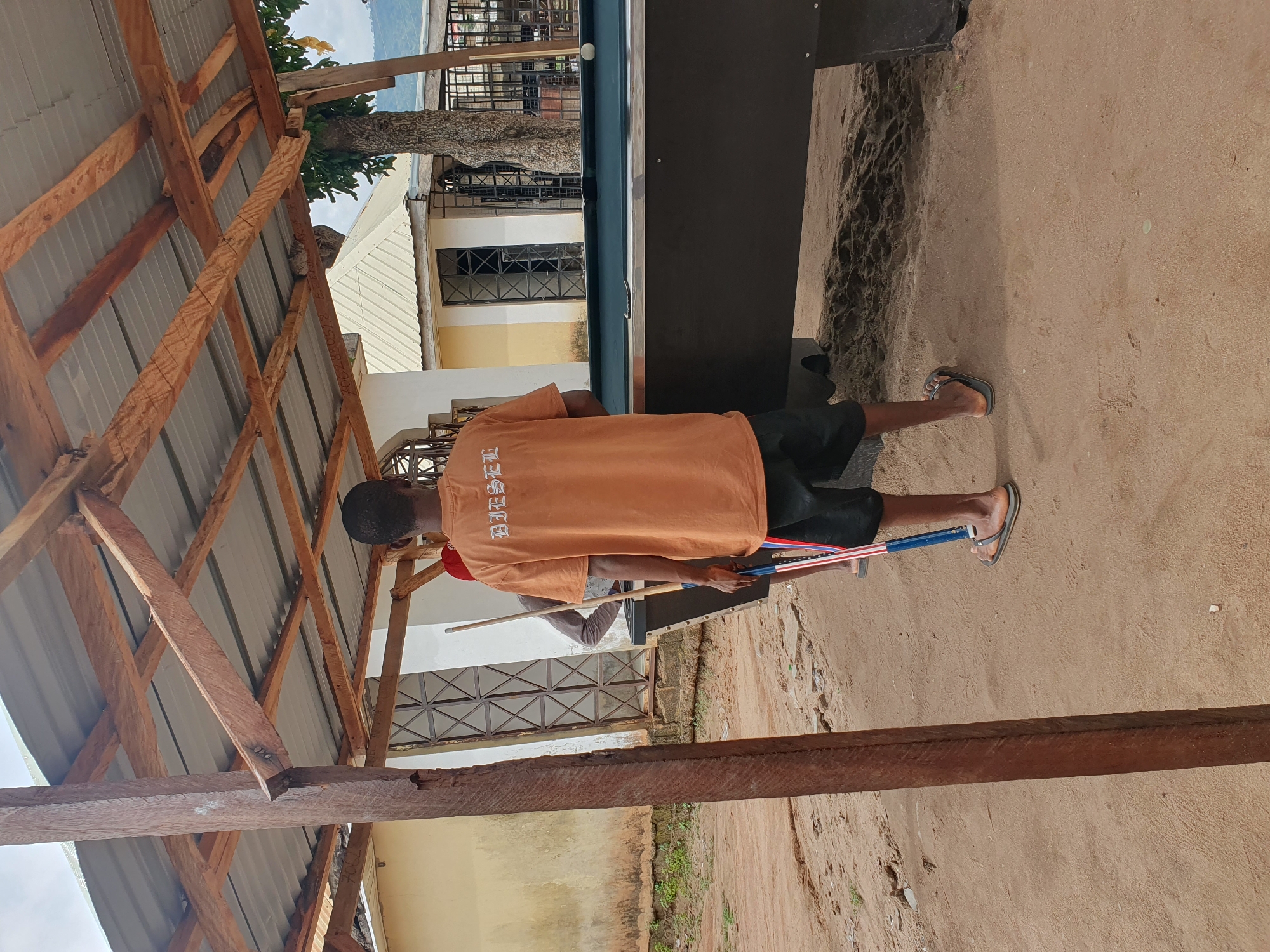 A male subject stands alongside a pool table in Lokoja, Nigeria. He wears an orange t-shirt with white lettering, black shorts, and sandals. He appears to be holding pool cues. The pool table is positioned outdoors on a sandy surface, partially shaded by a wooden-framed roof structure. A building with windows and a barred gate is visible in the background behind the pool table.
