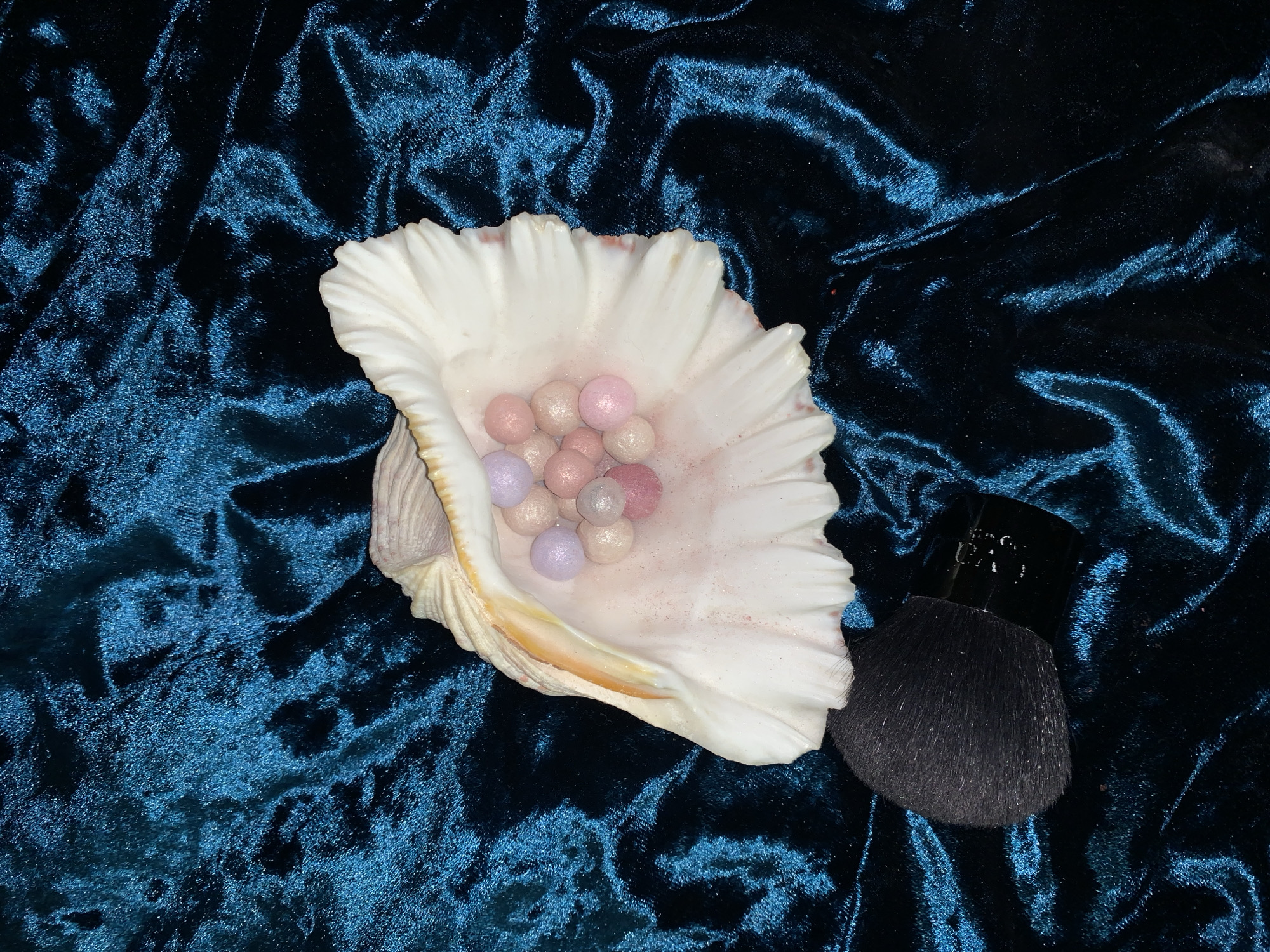 This media file displays a still life arrangement of cosmetic products on a dark teal velvet fabric. The primary subjects are a large, open seashell, filled with a collection of small, iridescent pearl-like makeup spheres, and a black makeup brush with a compact handle. The makeup spheres are a mix of pastel colors, including pink, light purple, and peachy tones, and they appear to have a shimmery finish. The seashell is white with hints of pink and orange along its ruffled edge, and it rests on the velvet surface, creating a natural cradle for the makeup pearls. To the right of the seashell, the black makeup brush is positioned with its bristles fanned out, suggesting it's ready for use or has just been used. The velvet fabric exhibits a rich texture with deep folds and highlights, indicating it is likely a plush material. The lighting in the image creates shadows and emphasizes the textures of both the fabric and the objects. There is no visible text in the image. The overall scene evokes a sense of elegance and a focus on beauty products, possibly arranged for photography or display. The setting appears to be indoors, with no discernible cues for time of day or weather. The mood is calm and sophisticated.