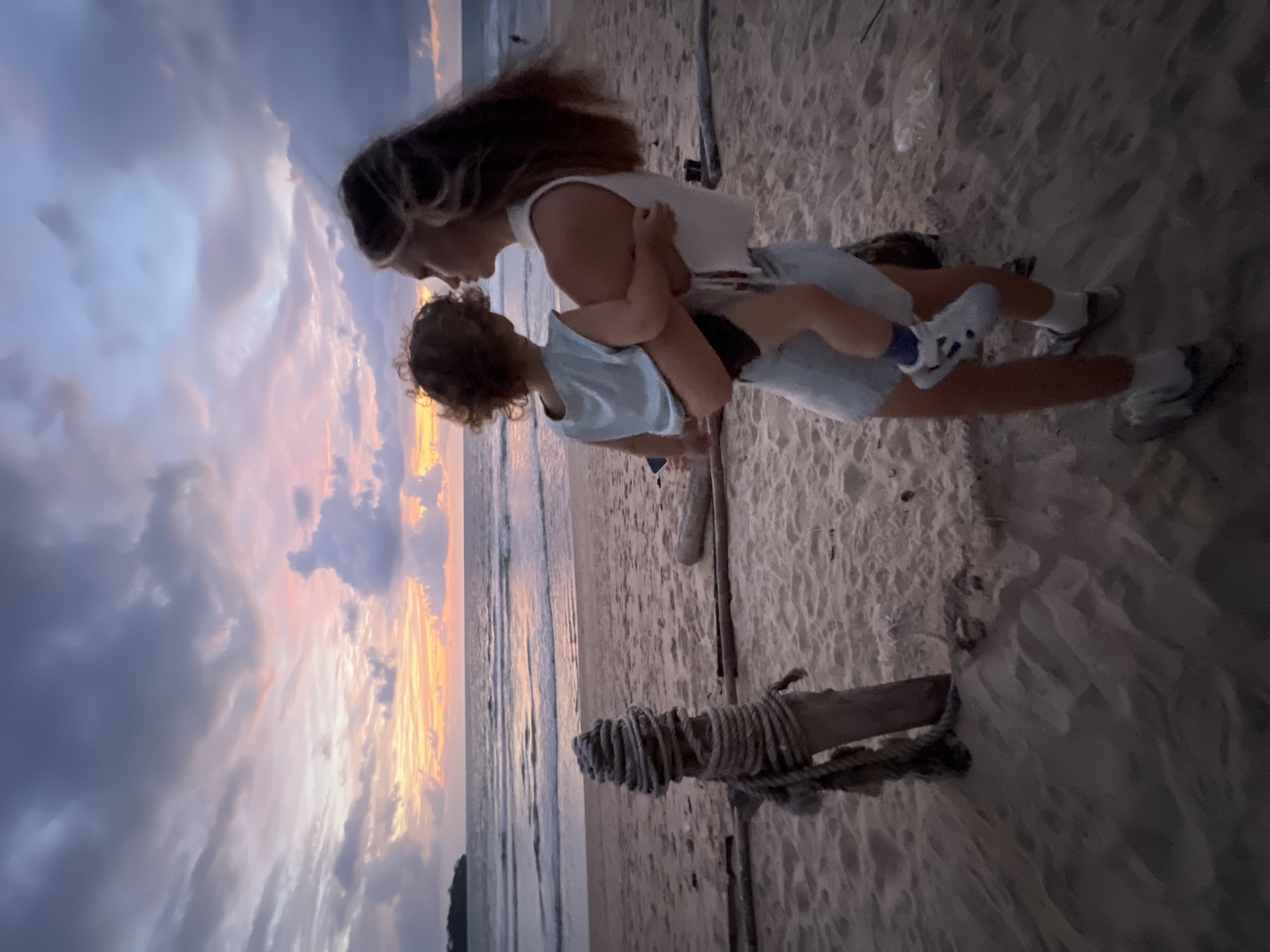 This media file captures a tender moment between a woman and a child on a beach at sunset. The subjects are a woman, likely a mother, holding a young child. They are positioned in the center of the frame, leaning towards each other, with the woman looking down at the child and the child looking up at the woman. They appear to be sharing an intimate moment, perhaps a hug or a playful interaction.
The setting is a beach, with sand visible in the foreground and middle ground. The ocean and waves are present in the background, suggesting a coastal location. The sky is filled with dramatic clouds, illuminated by the setting sun, casting warm hues of orange, pink, and purple across the scene. This indicates that the time of day is either sunset or twilight. The overall mood conveyed by the image is one of love, connection, and serenity, amplified by the beautiful natural lighting.
There are a few objects visible in the scene, including a piece of driftwood or a post with rope tied around it in the foreground, which adds a rustic element to the beach setting. The woman is wearing a white tank top and denim shorts, while the child is wearing a light blue t-shirt and shorts. Both are wearing sneakers.
The image appears to be taken in Choeng Thale, Thailand, as per the location context. There is no visible text within the image itself. The image is oriented vertically, with the beach and ocean running along the left side of the frame and the sky dominating the upper portion.