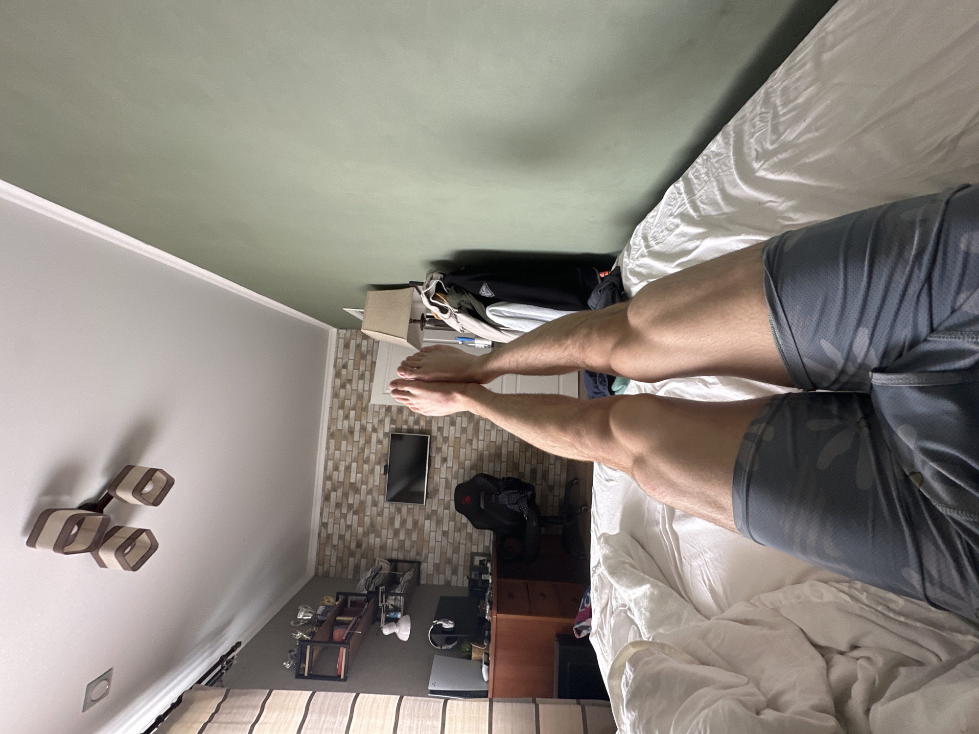 The scene depicts a first-person view of a person lying on a bed in a bedroom. The person is wearing grey camo shorts and has their legs extended towards the end of the bed. The bed is covered with a white comforter. The bedroom has a green-painted ceiling and grey walls. On the wall is a geometric-shaped light fixture. Adjacent to the bed is a wall featuring a decorative brick pattern. A television is mounted on this wall above a desk. On the desk, there are computer peripherals, including a headset and a PlayStation 5 gaming console, along with a black gaming chair. There are some shelves with various items on them in the background. The location is in an unknown city.