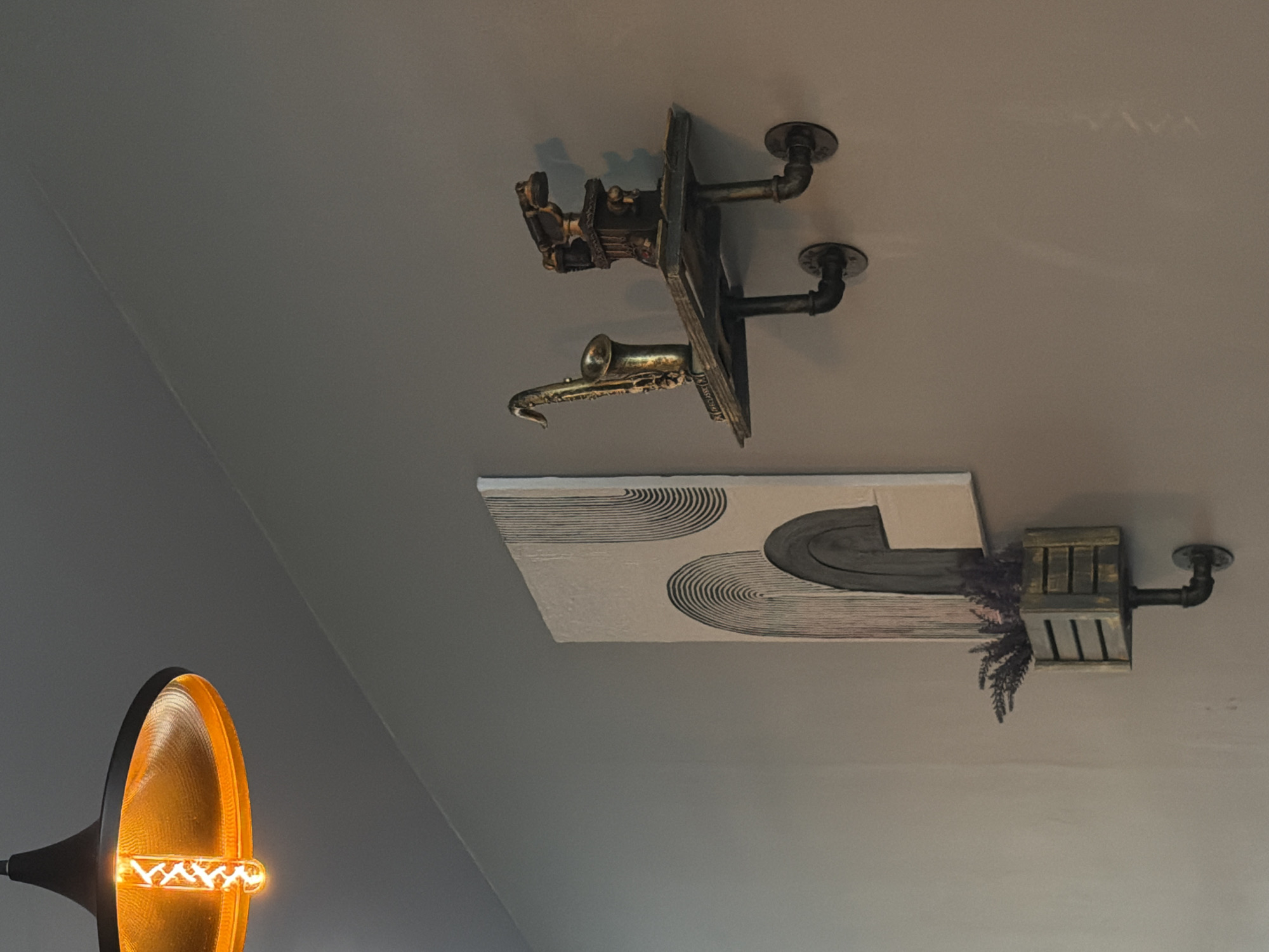 The image depicts a portion of a room's ceiling with decorative elements. In the upper center is a mounted art piece featuring a golden saxophone and what appears to be an antique radio or phonograph, both attached to a wooden plaque with iron pipe mounts. Below this, a rectangular canvas displays black semi-circular patterns next to a raised, curved element reminiscent of a road, which leads into a wooden crate with purple lavender sprouting from it. This is also mounted with iron pipe. In the lower left corner, the warm glow of an exposed filament bulb is visible within a black metal lampshade. The setting appears to be indoors, with the ceiling painted a neutral gray. There is no information on whether the image was taken in Birobidzhan, Russia.