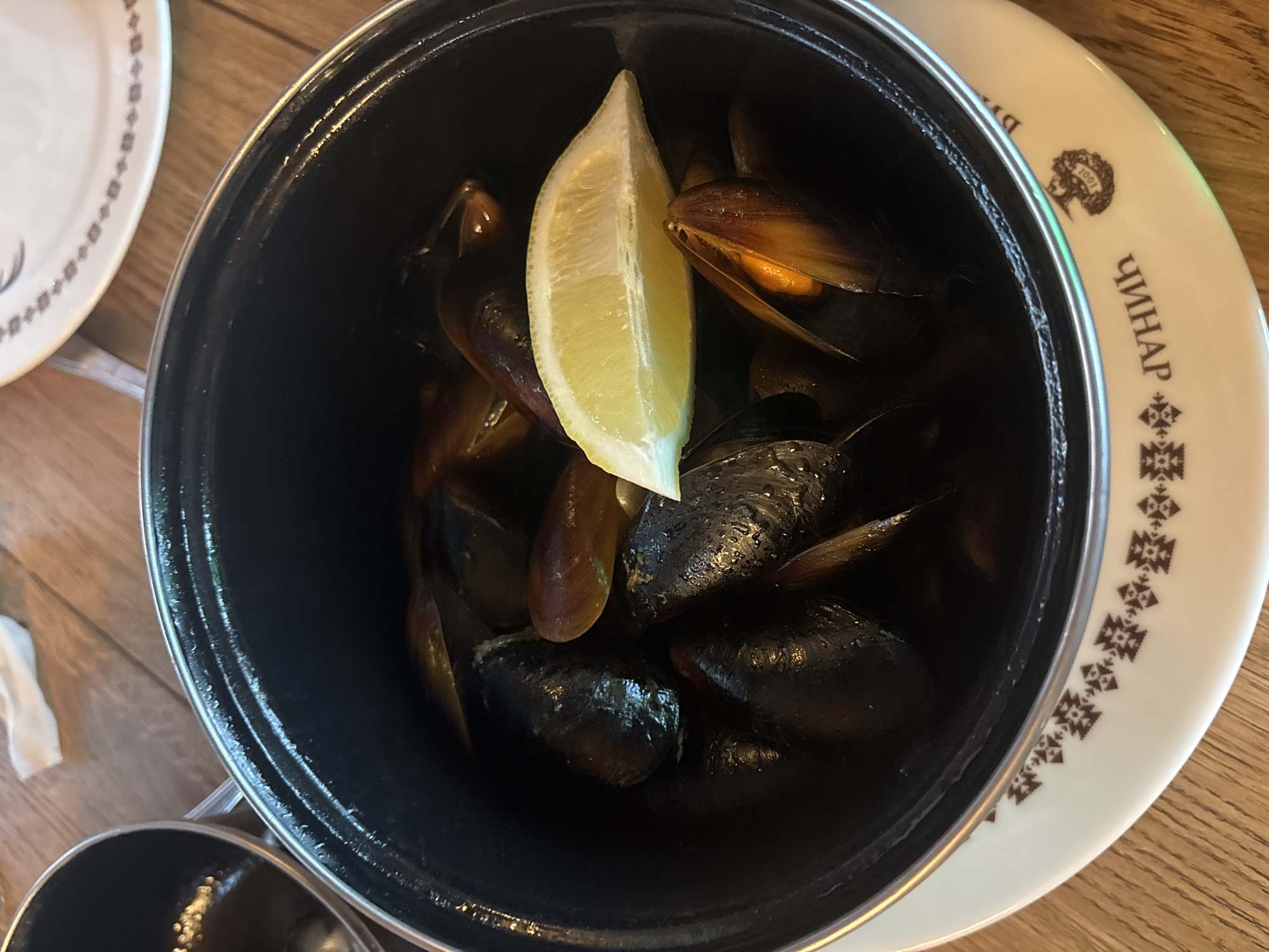 A top-down shot reveals a black pot filled with mussels and a lemon wedge, resting on a white plate with brown decorative markings and the word "ЧИНАР" (Chinar) in Cyrillic, indicating a restaurant name. The plate sits on a wooden table. The location is likely Sofia, Bulgaria, based on the Cyrillic inscription.