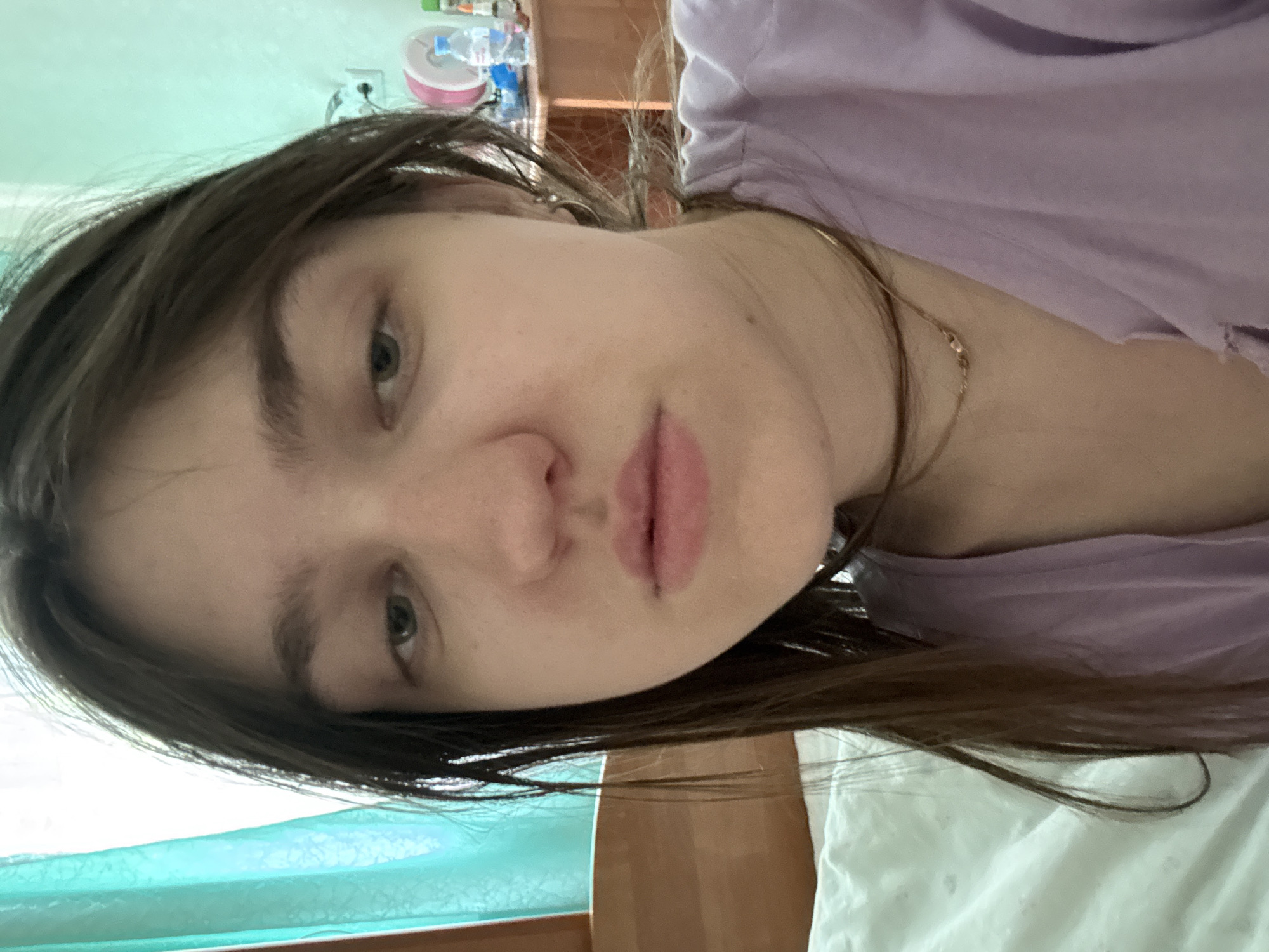 A close-up, rotated selfie shows a fair-skinned person with dark brown hair and green eyes. The person is wearing a light purple garment. A thin gold chain is visible around the neck. The background includes blurred elements like a light-colored wall, a window with a light blue hue, and some household items. The location is not identifiable from the image.