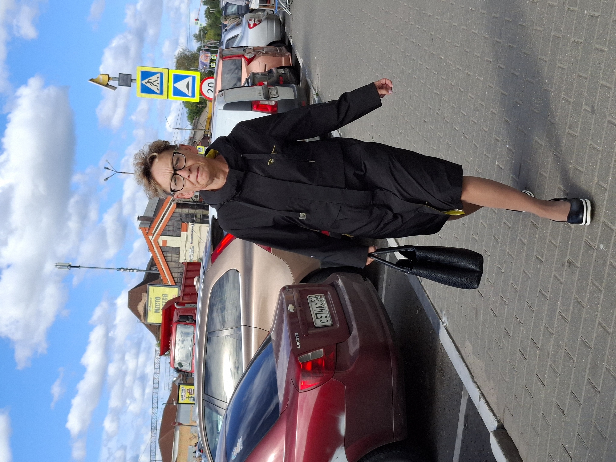 An adult woman with short, styled, graying hair and glasses walks on a paved walkway beside a parking lot. She is wearing a black jacket and a black skirt, with yellow trim visible on the inside of the jacket collar. She carries a black, textured handbag in her right hand. In the background, several cars are parked, including a red Chevrolet Lacetti with a license plate reading "C574AC 50". Beyond the cars, buildings with orange and white trim are visible, along with a red dump truck and traffic signs. The sky is blue with white, fluffy clouds.