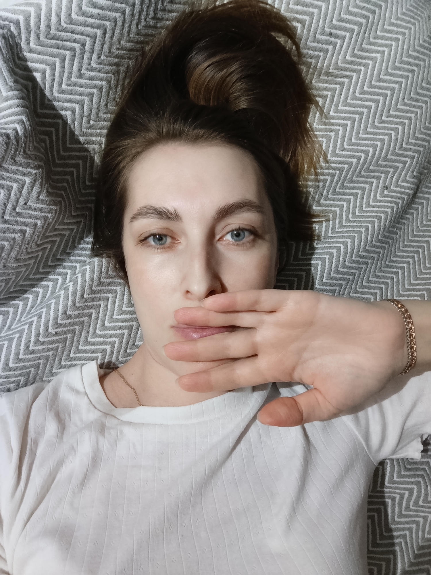A fair-skinned person with light brown hair piled loosely on top of their head is lying on a gray and white patterned fabric, possibly a bedspread or blanket. The person is wearing a white shirt with a subtle textured pattern. They are positioned to face the camera directly, with their right hand covering the lower half of their face, up to the nose. A gold-colored chain bracelet is visible on their right wrist. The person has blue eyes and appears to be without makeup. A gold-colored chain necklace is visible at the base of their neck. The location is not discernible from the image.