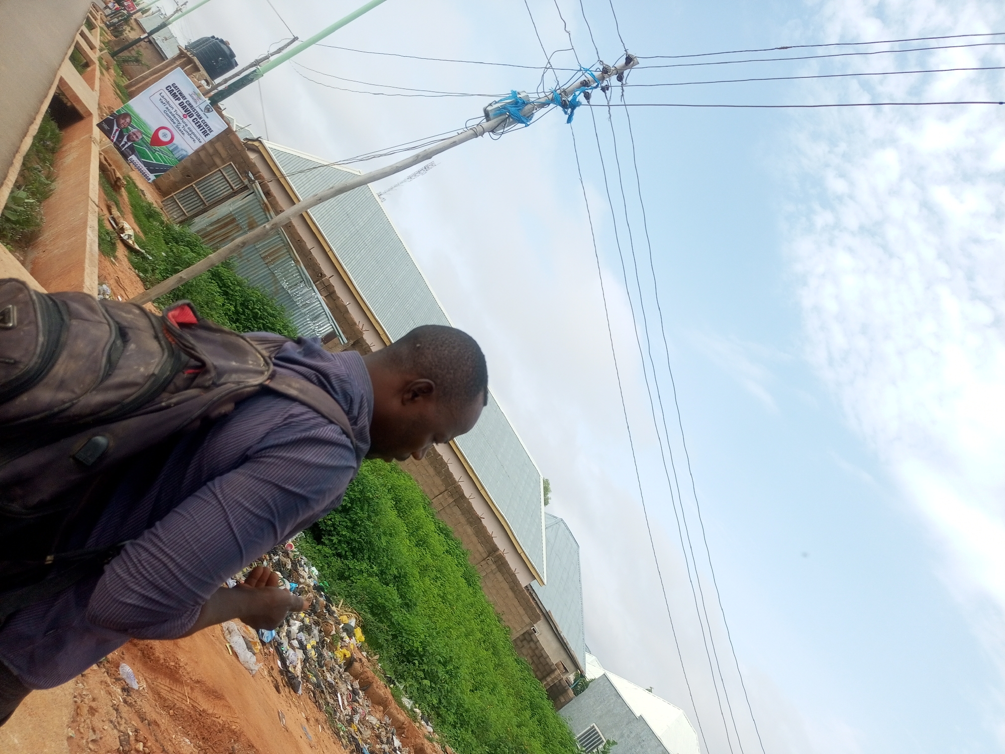 A man wearing a purple striped shirt and a backpack is walking next to a pile of garbage. The man is facing to the right, looking down. The garbage is on the side of a dirt road, with green vegetation growing behind it. In the background, there are several buildings with metal roofs and a billboard advertising "Gateway Christian Centre Camp David Centre." Power lines are visible overhead against a pale blue sky with some clouds. The image appears to have been taken in Gombe, Nigeria.