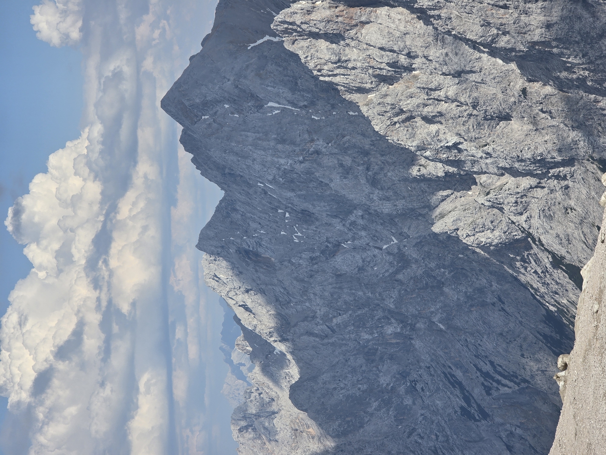 The image depicts a vast mountain range under a cloudy sky. The mountains are primarily composed of gray rock with visible textures and shadows indicating their rugged terrain. Patches of snow are scattered across the peaks and crevices. In the foreground, the ground is covered with gravel and small stones. The sky is blue with large, white cumulus clouds and some lighter, more scattered clouds. The scene appears to be a natural, mountainous landscape. The query specifically asks for a location in Garmisch-Partenkirchen, Germany. The Zugspitze, Germany's highest peak, is located near Garmisch-Partenkirchen and its landscape often features similar mountain formations and cloudy skies. Therefore, it is highly probable that this image was taken in the vicinity of the Zugspitze. There are no people or specific objects depicted that would indicate human activity or a city environment.