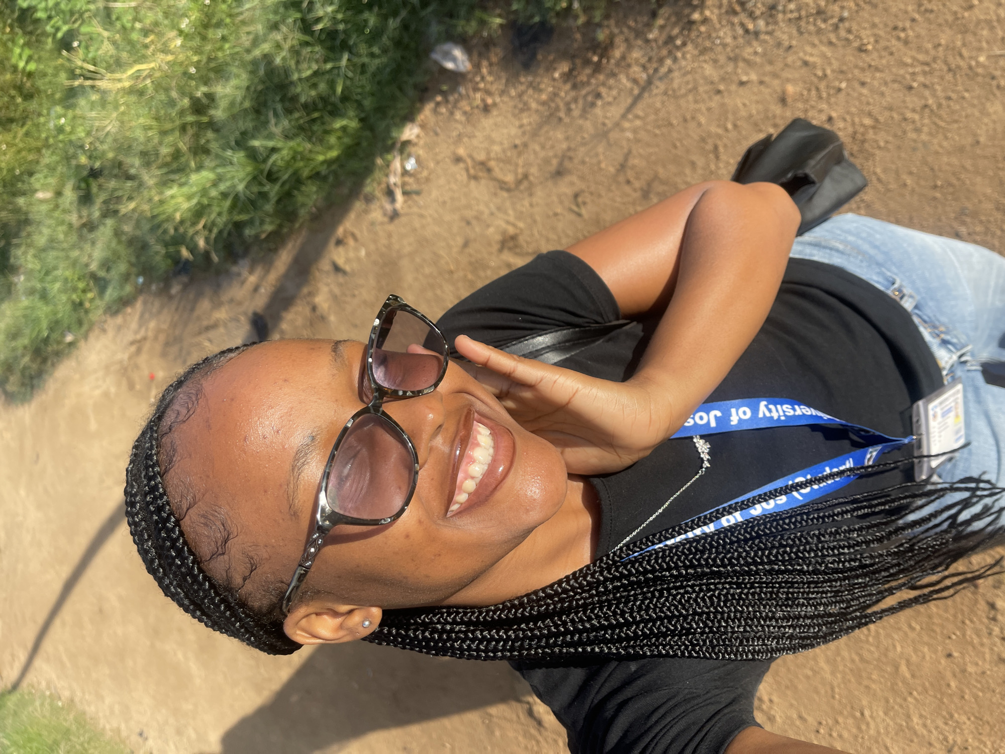 In a selfie taken outdoors, a young Black woman with braided hair and sunglasses smiles, her right hand touching her cheek. She wears a black t-shirt, blue jeans, and a lanyard with a University of Jos ID. The background shows a dirt path and green foliage, consistent with an outdoor setting in Rafin Par, Nigeria.