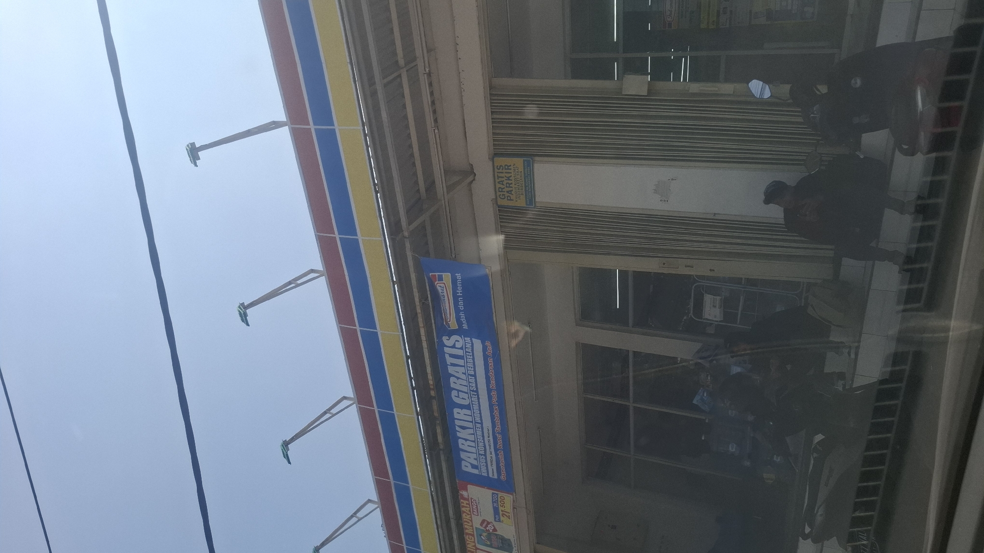 The image depicts the exterior of an Indomaret convenience store, a common chain in Indonesia, under a bright, clear sky. The store features a red, blue, and yellow striped awning. Above the awning, several metal poles with green tops extend upwards, likely for lighting or decorative purposes. A prominent blue banner above the entrance reads "PARKIR GRATIS KHUSUS KONSUMEN INDOMARET SAAT BERBELANJA" which translates to "FREE PARKING FOR INDOMARET CUSTOMERS WHILE SHOPPING." Below this, smaller text details that the free parking is for customers who purchase something and mentions "Jumlahkan Kunci Tambahan Pada Kendaraan Anda" meaning "Sum the Additional Keys on Your Vehicle." To the right of the main entrance, a smaller yellow sign also states "GRATIS PARKIR" with similar conditions.
In front of the store, several people are visible. Two individuals are sitting on a raised platform near the entrance, one wearing a baseball cap and looking at their phone, while another person is obscured by darkness. A motorcycle is parked to the left. The lower part of the image shows a reflection, likely from a car window, overlaying the scene with a dark, grainy filter. The overall environment suggests a sunny day in an urban or semi-urban area, characteristic of a location like Rancagoong, Indonesia, where such convenience stores are prevalent.